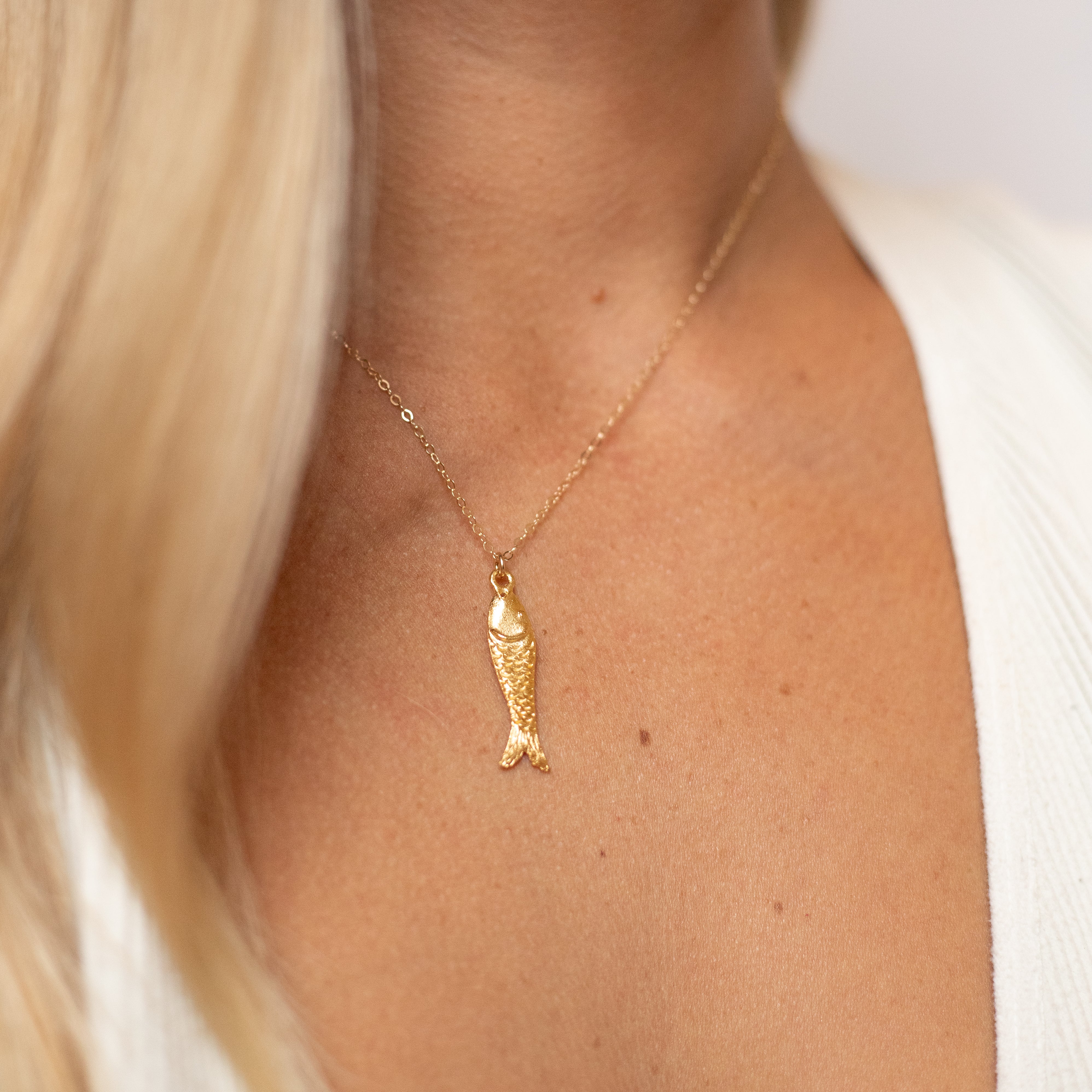 One inch long gold fish charm on a gold chain, shown on a woman's neck. 