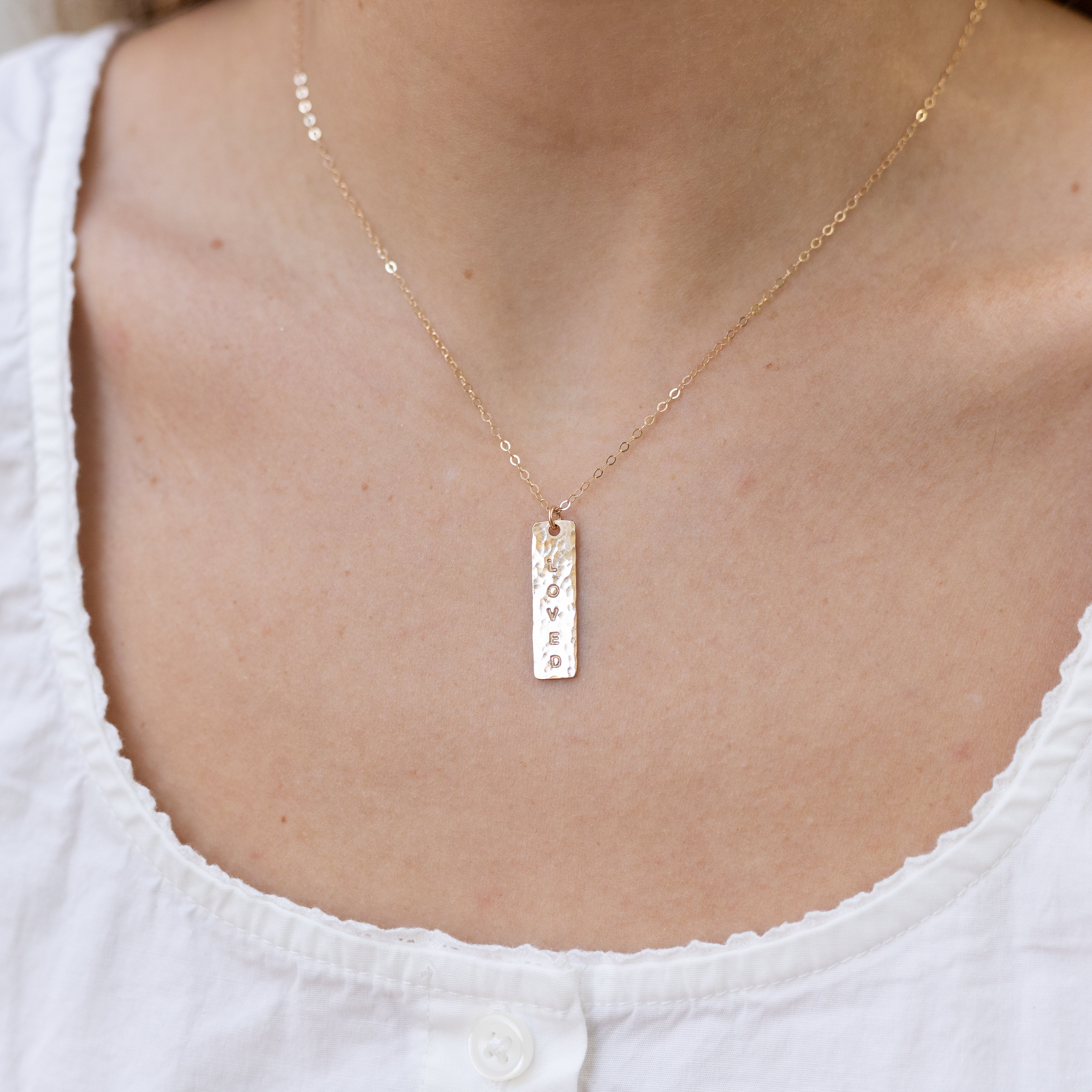 About an inch long vertical gold bar with “Loved” stamped onto it. On our basic gold chain. Shown on a woman’s neck.