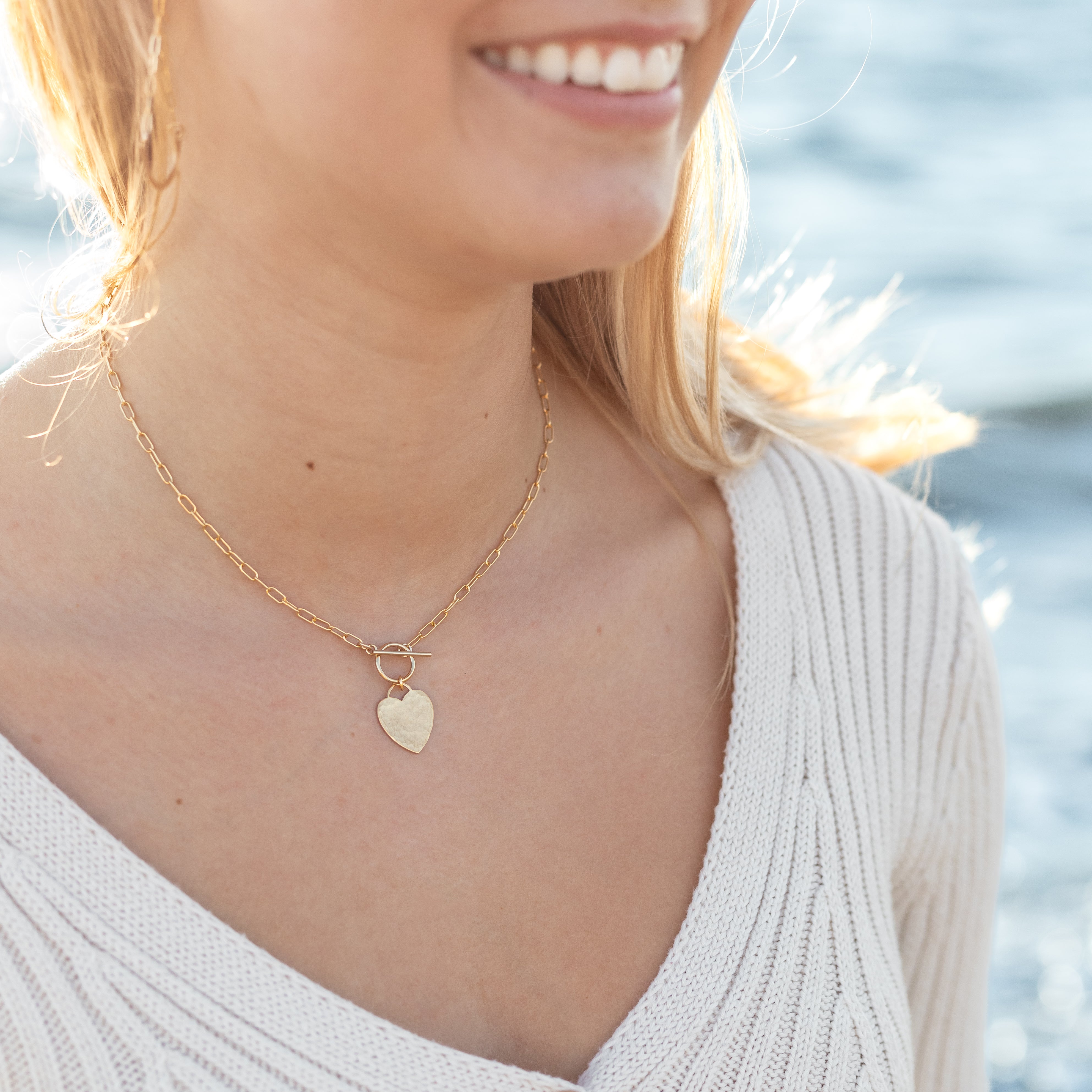Gold filled heart hammered and hanging from a toggle clasp. Attached to paperclip chain. Shown on a woman’s neck.
