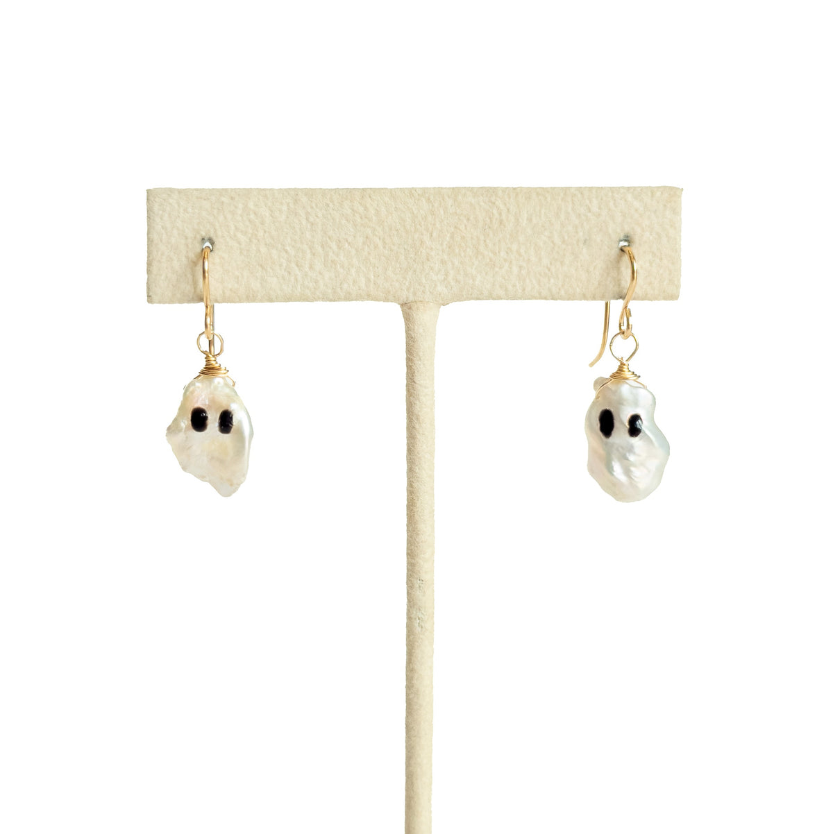 A pair of ghost-shaped pearl drop earrings, small black eyes, and gold hooks, displayed on a cream-colored jewelry stand against a white background.