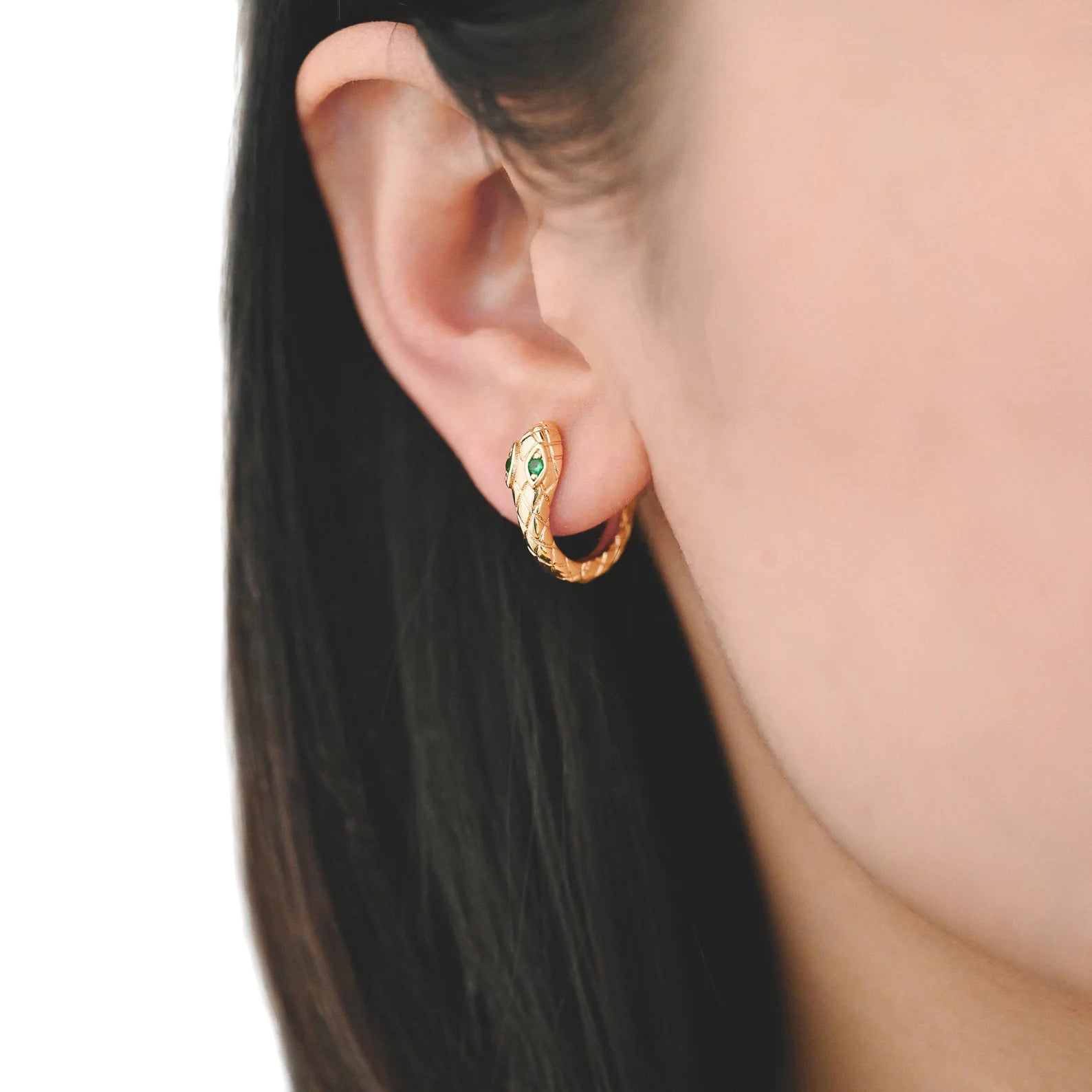 Gold plated snake huggie hoop earrings with green stones as the eyes. Shown in a woman’s ear.