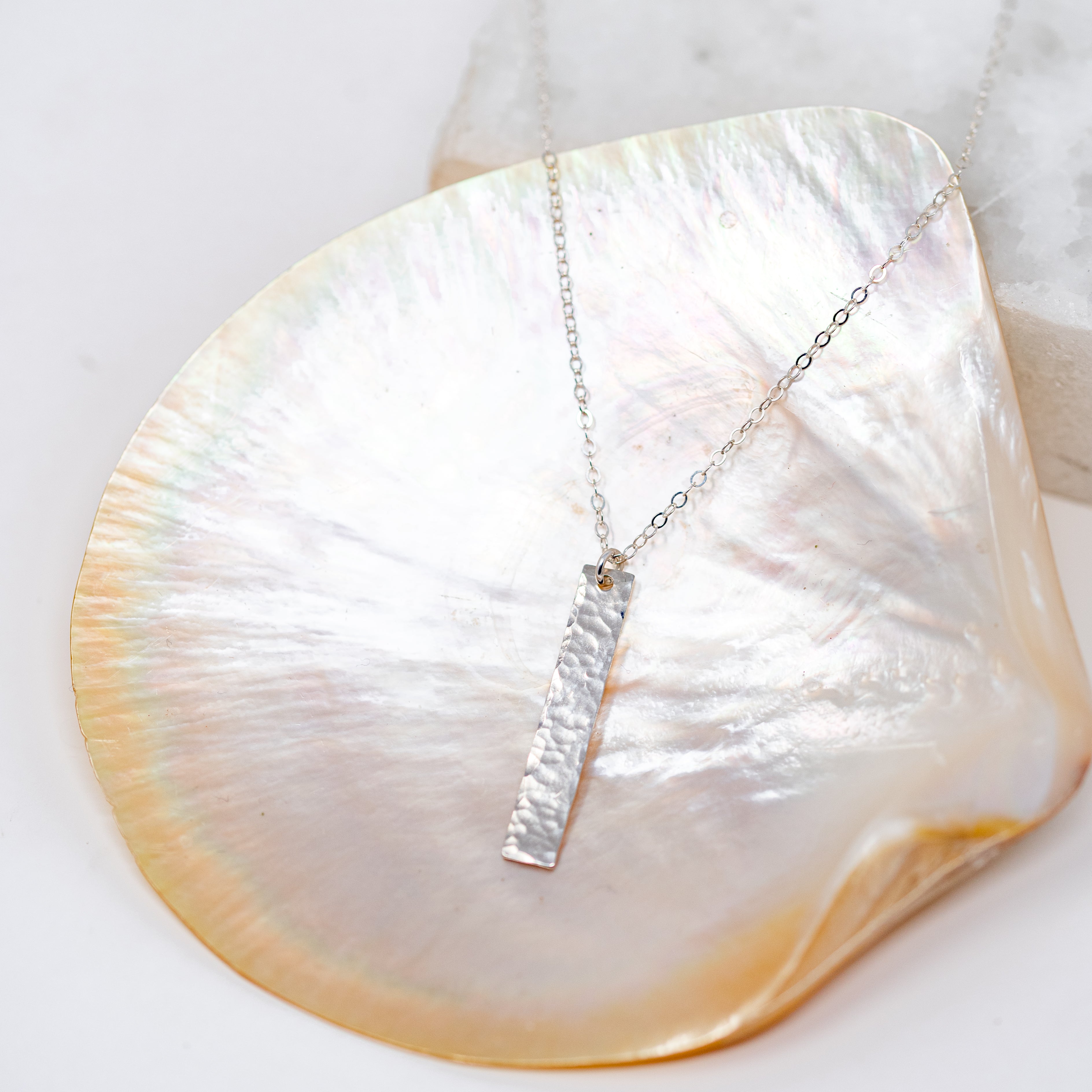 A silver vertical bar that is around an inch to an inch and a half. Attached to a silver chain and pictured in a shell. 