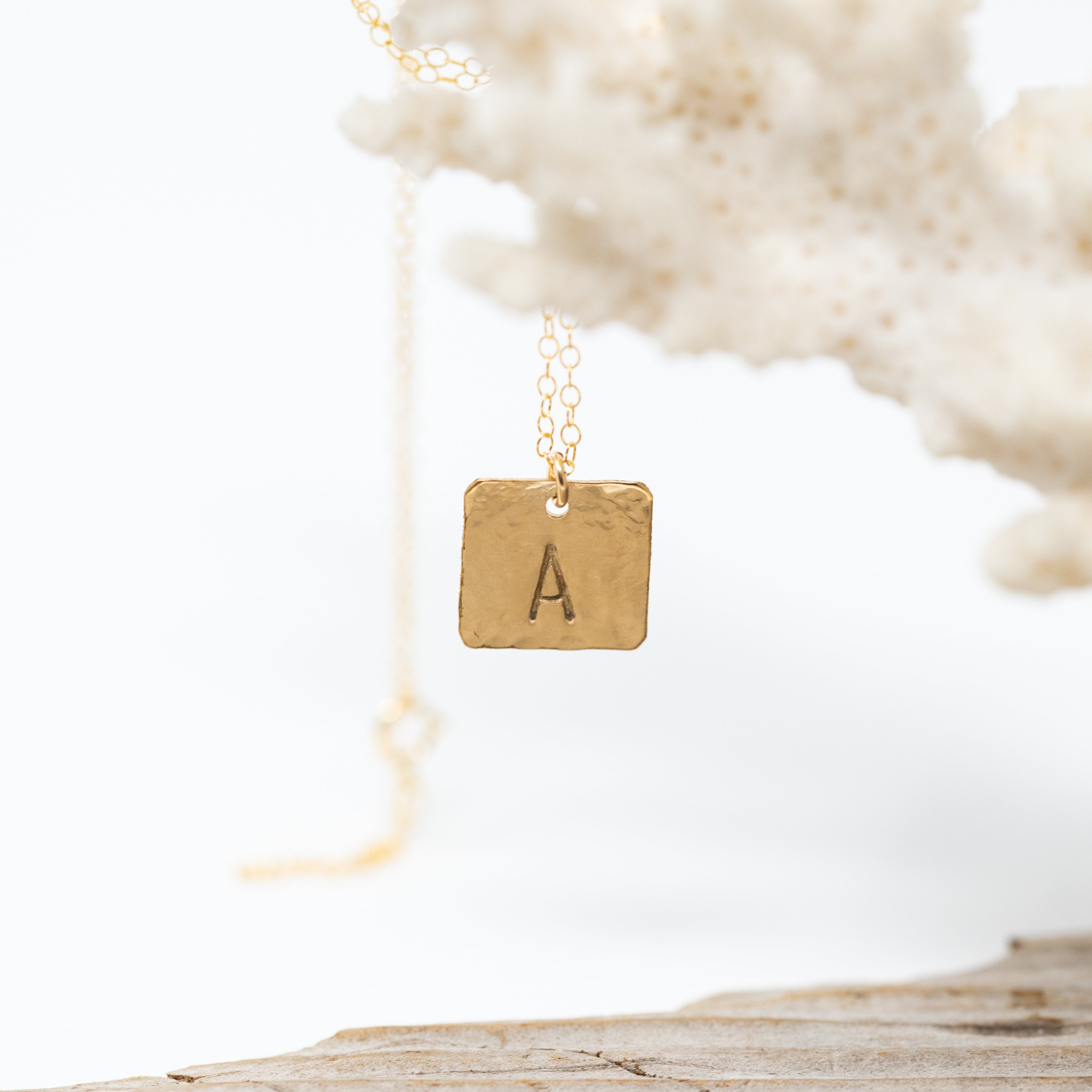 Small dime sized gold square with one large initial stamped onto it, attached to a chain. Hanging from a piece of coral. 