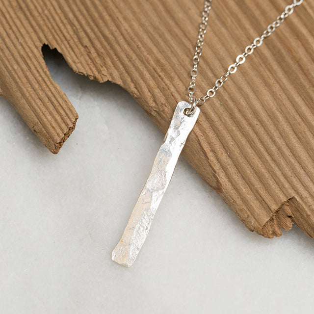 A silver vertical bar that is around an inch to an inch and a half. Attached to a silver chain and pictured on a piece of wood. .