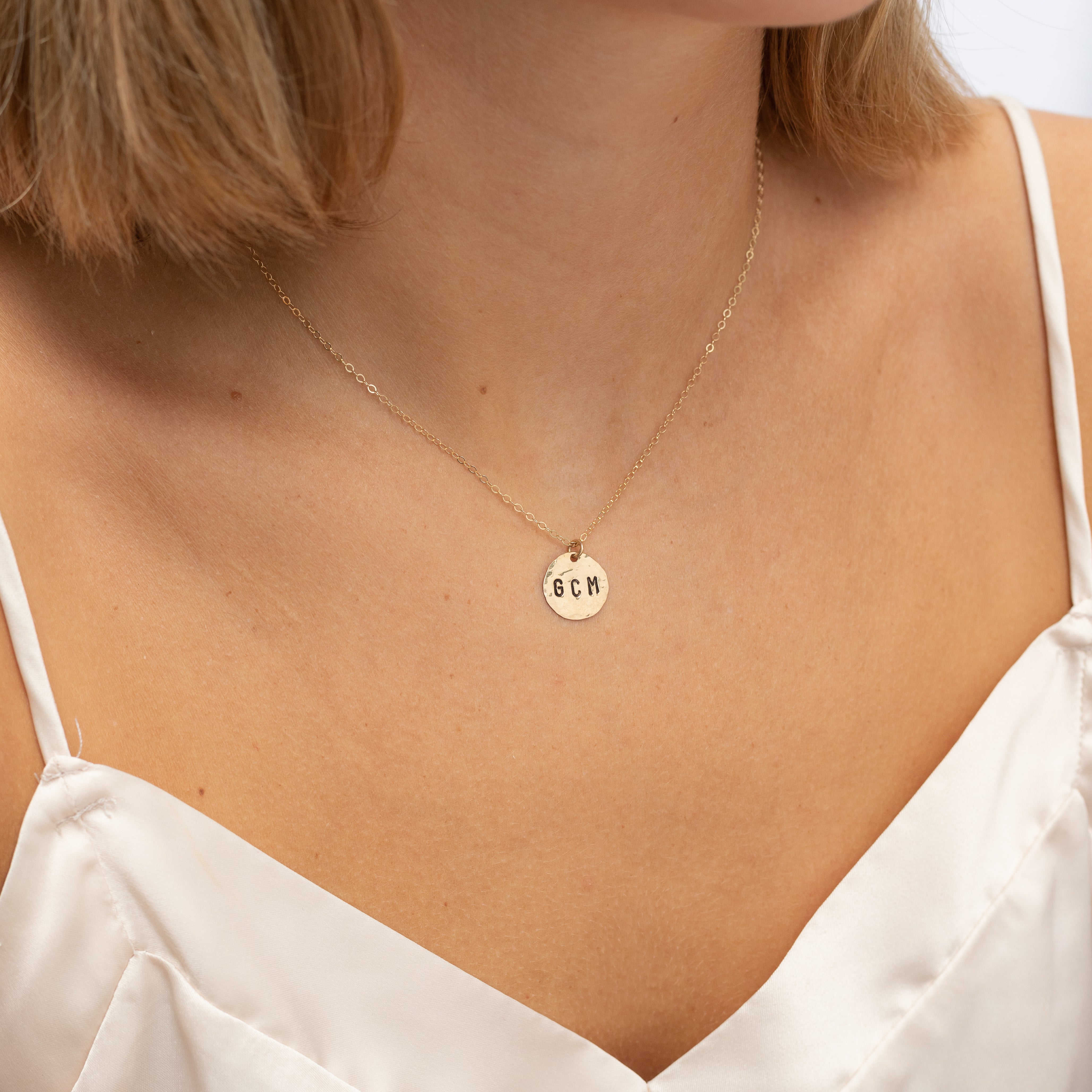Medium size gold filled hammered circle with initials stamped onto it. On a gold chain. Shown on a woman's neck. 