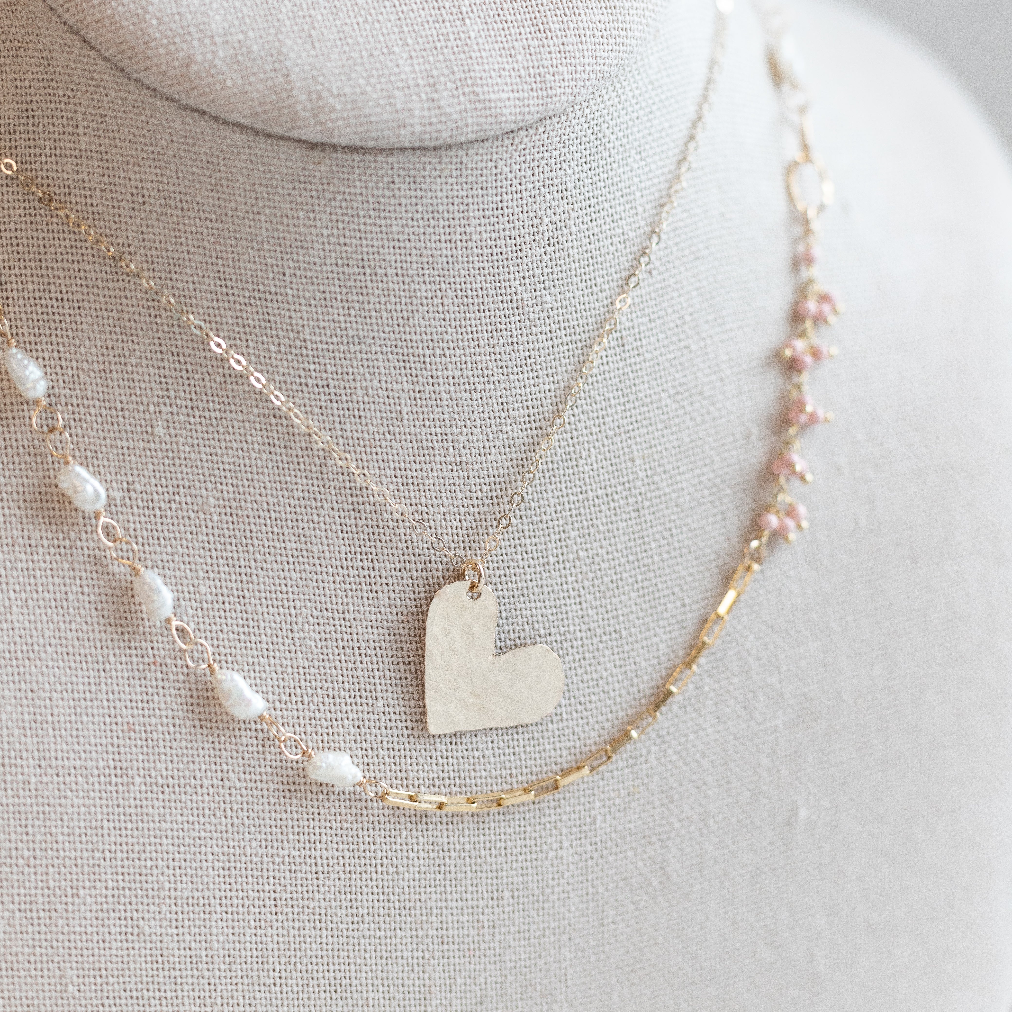 gold hammered hand cut heart charm nickle-quarter size, attached to a gold chain. Gold necklace below it with some pearls attached to it.