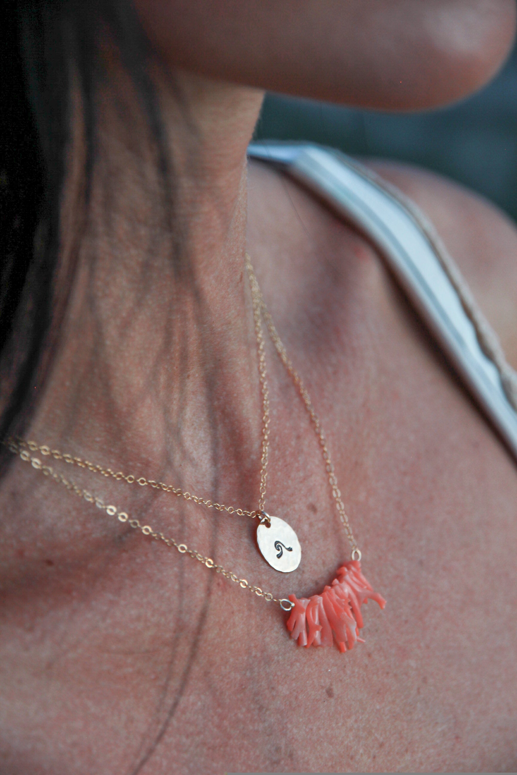Tiny gold filled circle with a wave stamped onto it. On a gold chain. shown on a woman's neck with another coral necklace below it. 