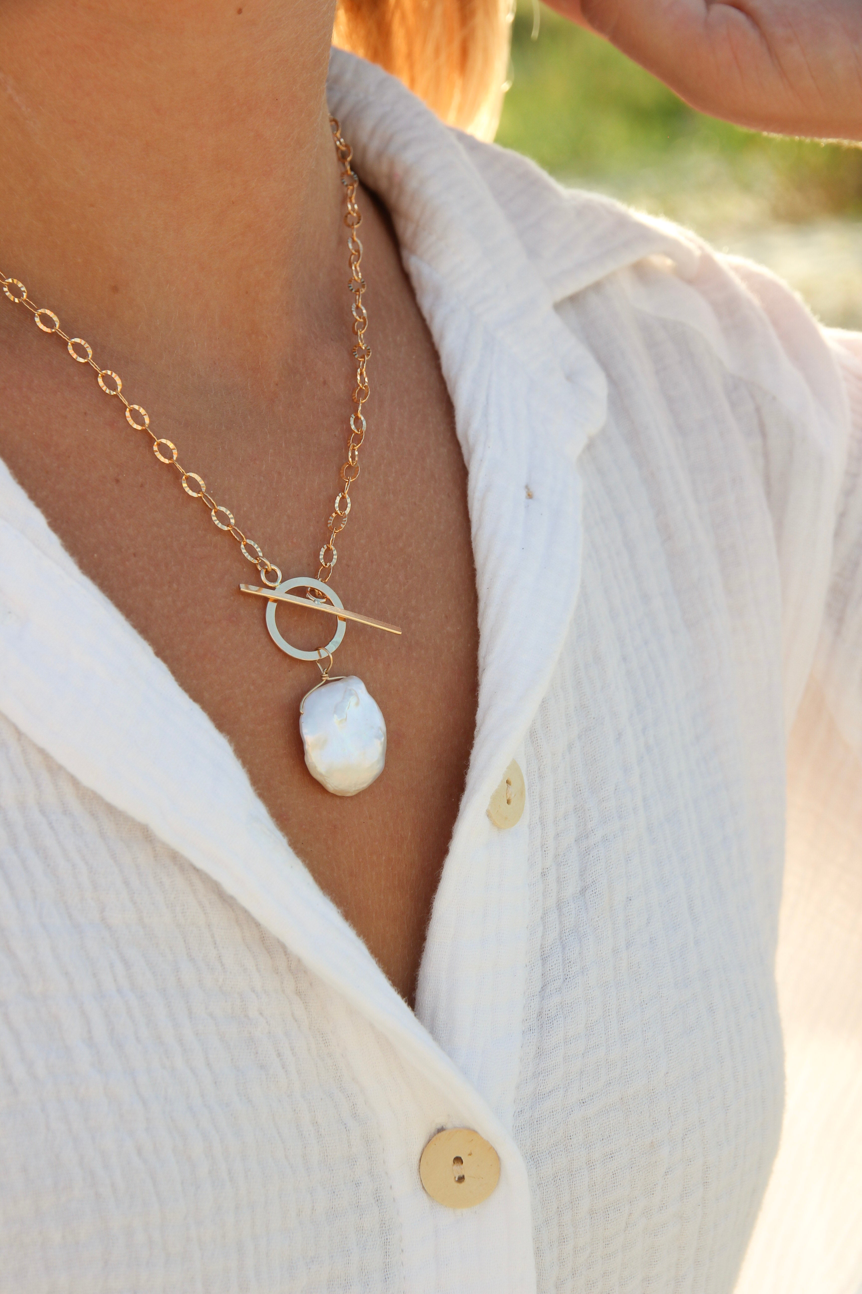 Gold chain necklace with a toggle clasp. There is a dime sized Cornflake pearl wire wrapped on the circular part of the clasp. Shown on a woman’s neck.