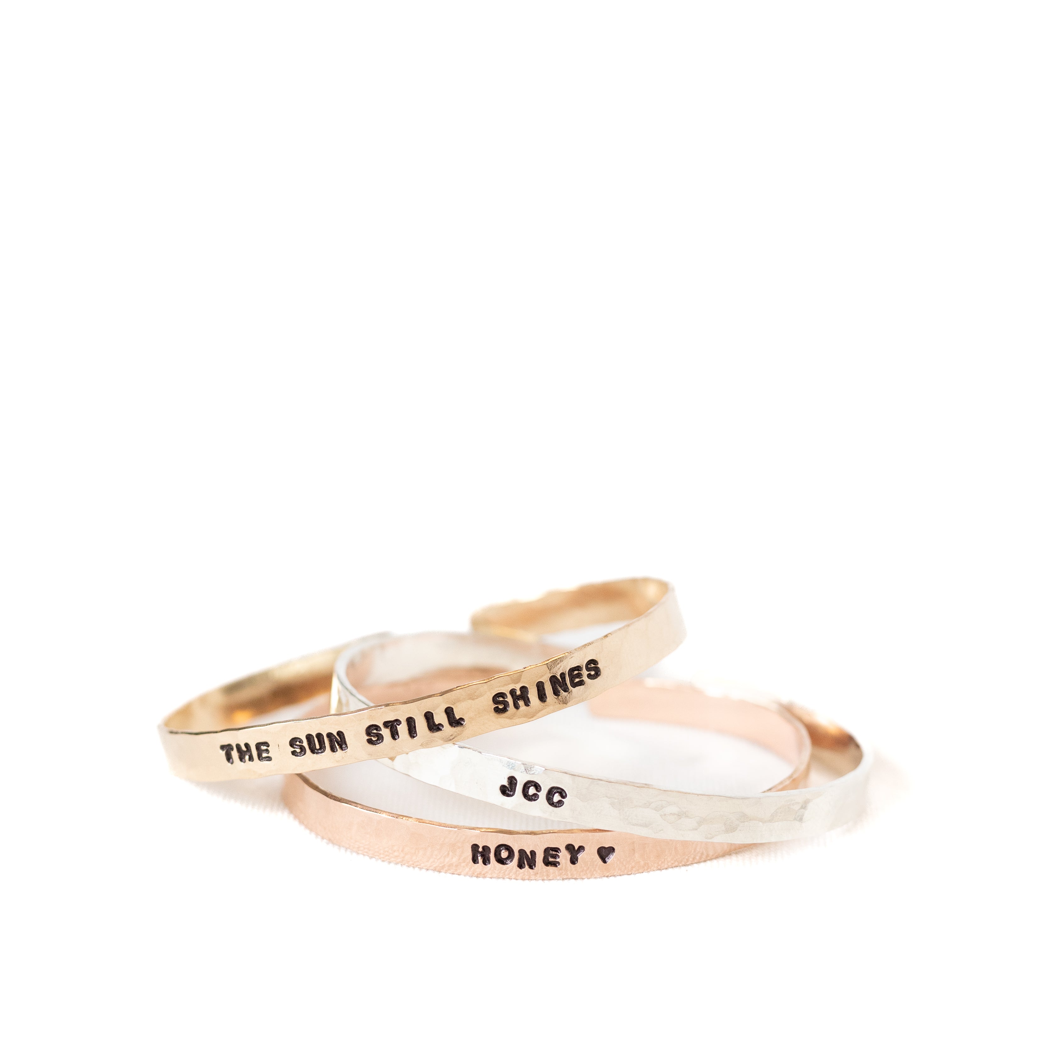 Three different cuffs with phrases and initials stamped on the outside of each cuff. There is a gold, sterling silver, and rose gold cuff pictured