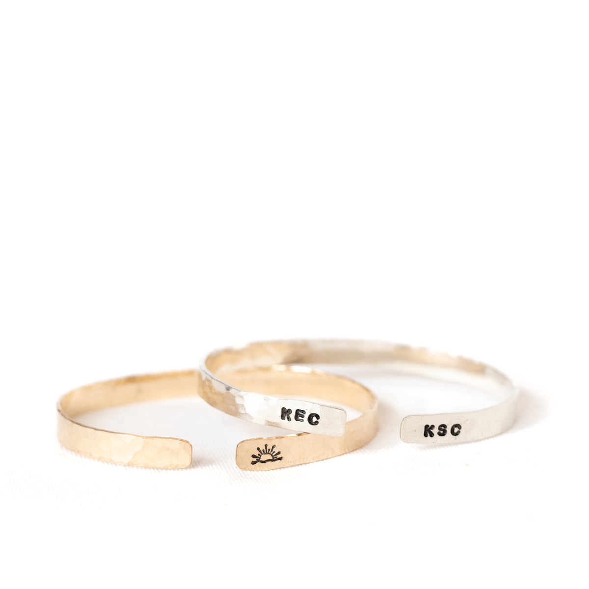 Two cuffs with symbols and initials stamped on the outside edge of them. The left one is gold with a sunset stamped and the right one is silver with initials stamped onto it.