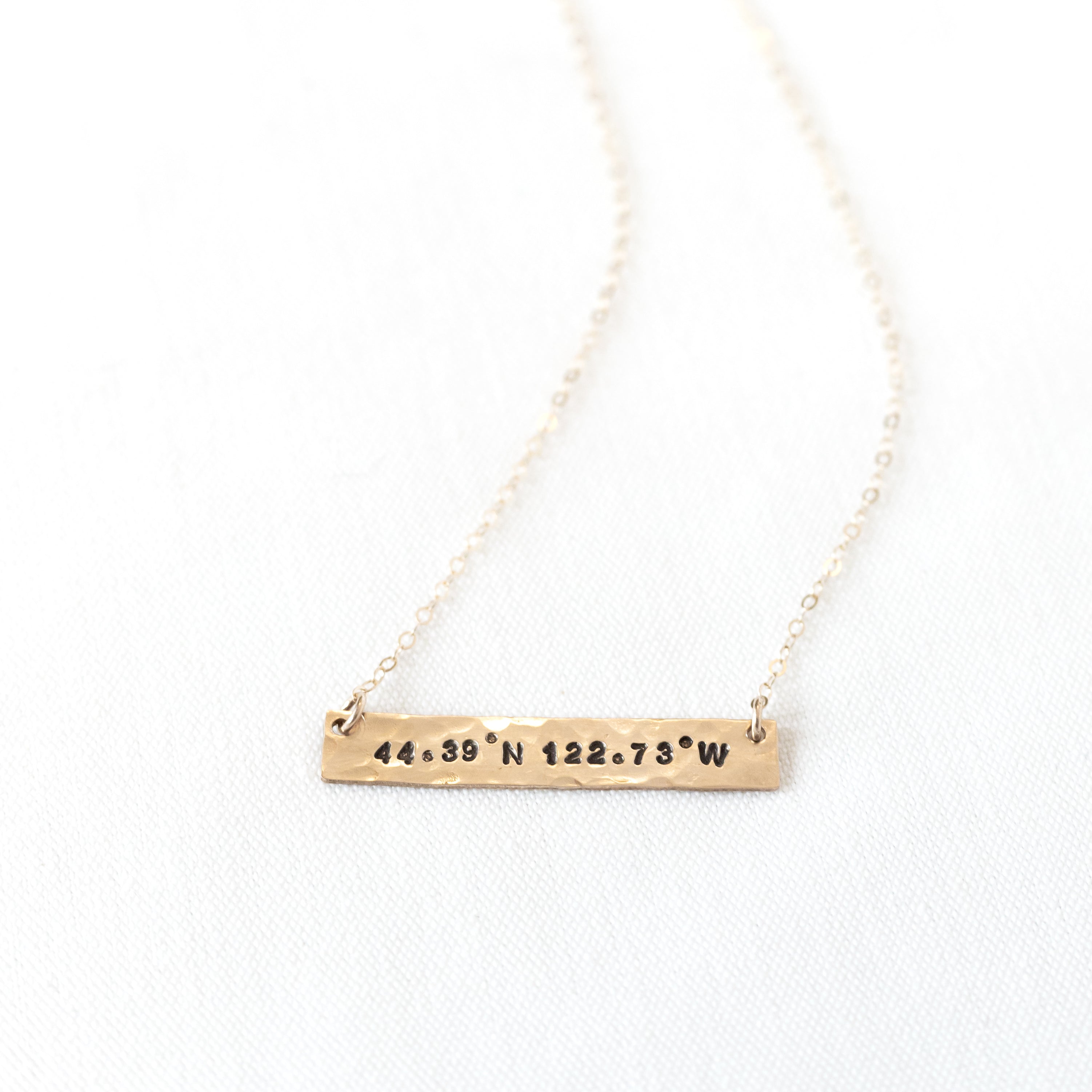 Horizontal gold bar around 1.5 inches long. Coordinates stamped on the bar and filled in with black coloring on a gold chain.