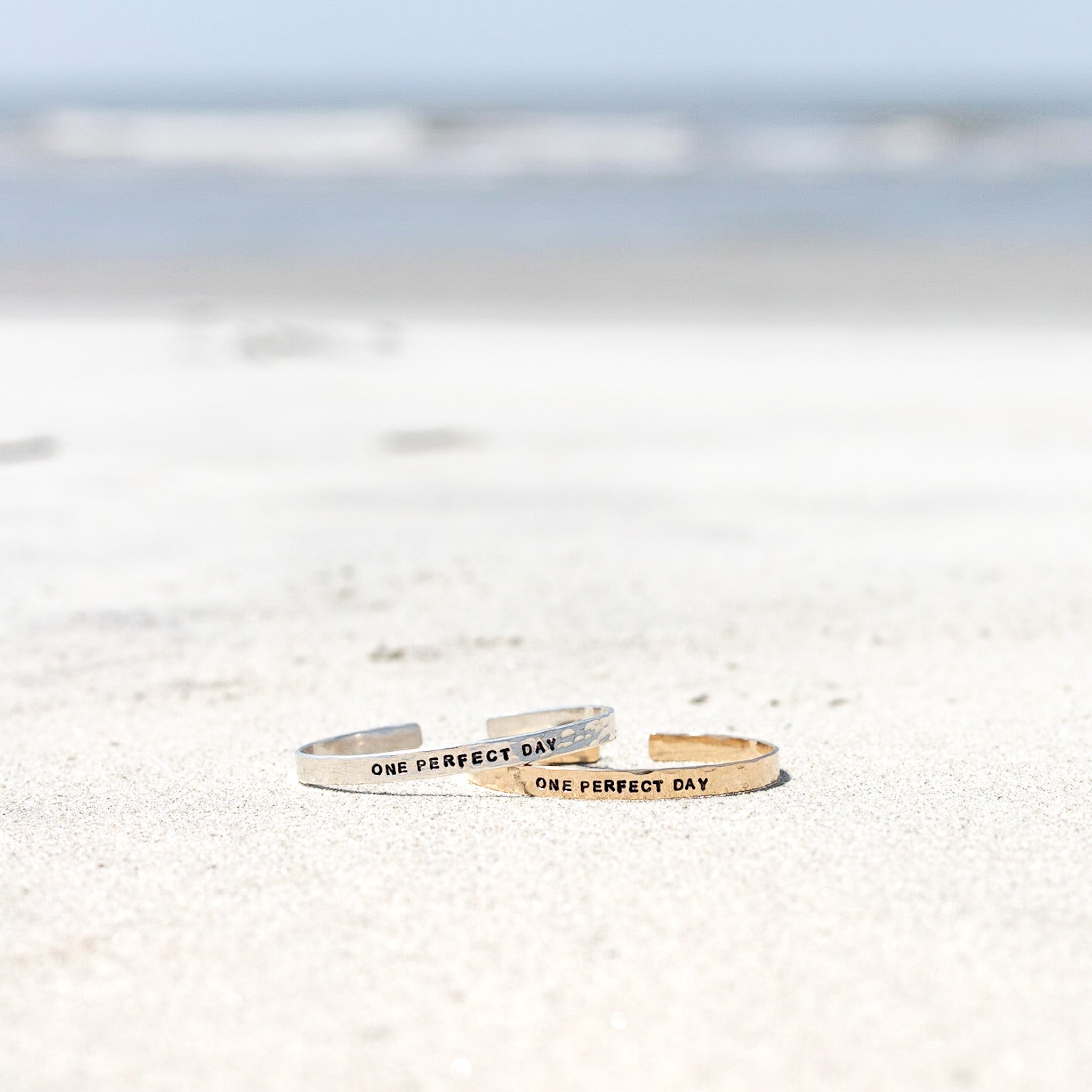A gold filled and sterling silver cuff with the phrase "one perfect day" on it. Shown on a beach. 