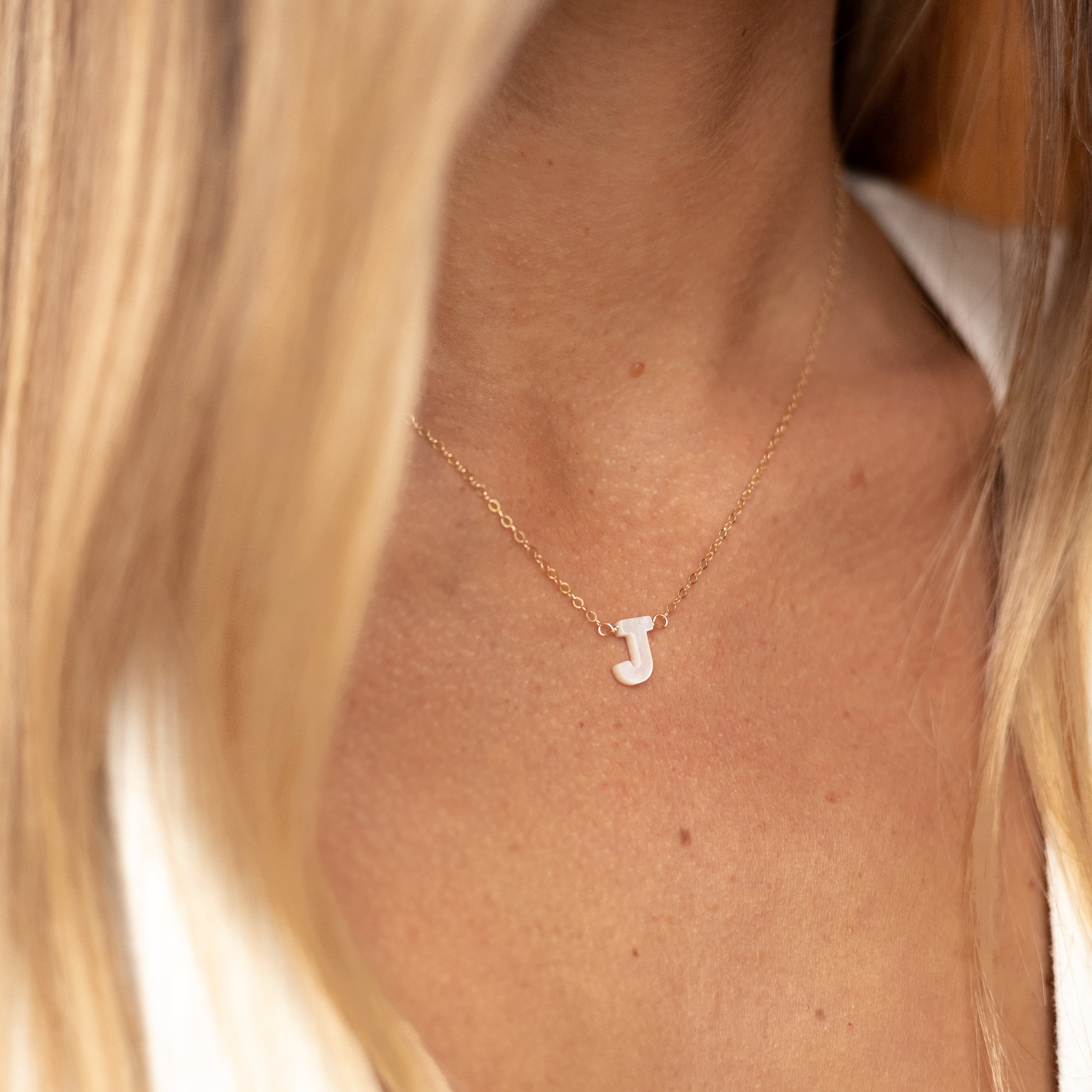 Mother of pearl initial "J" on a gold chain. Shown on a woman's neck. 
