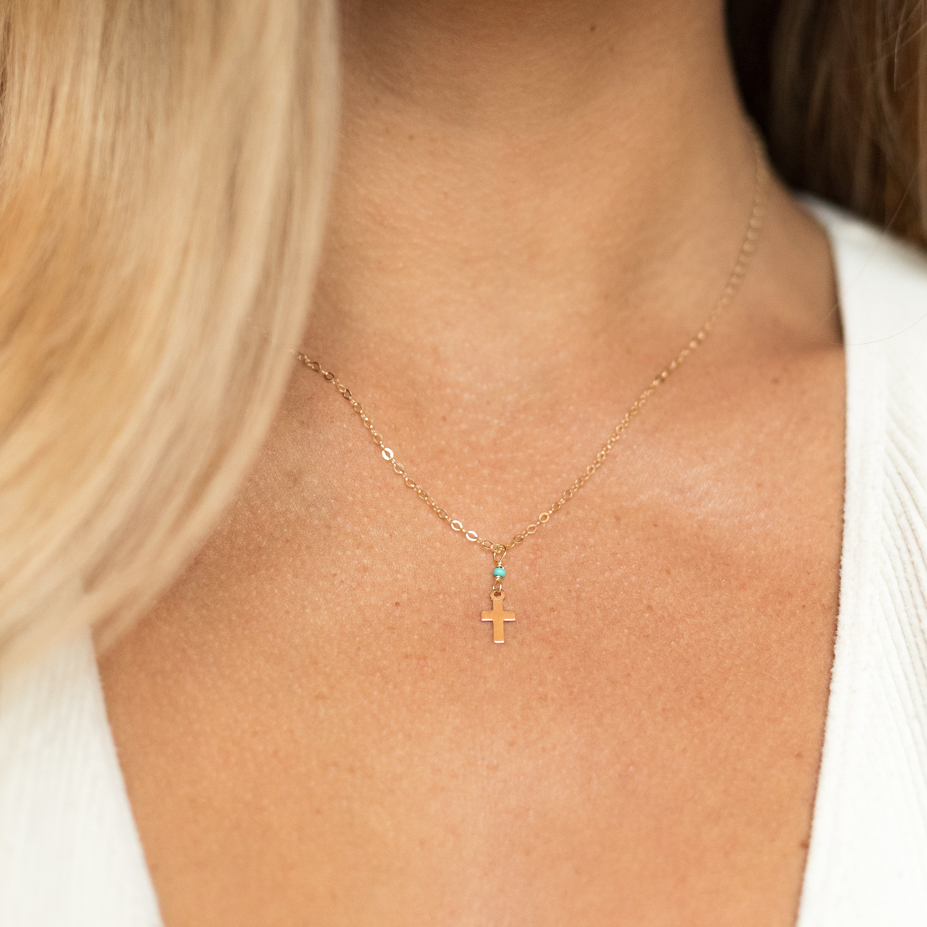 Tiny gold filled cross necklace with a tiny turquoise bead attaching it to a gold chain. Shown on a woman's neck. 