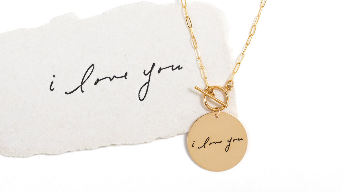 Gold necklace with 'I love you' pendant on a white card with the same text, on a white background