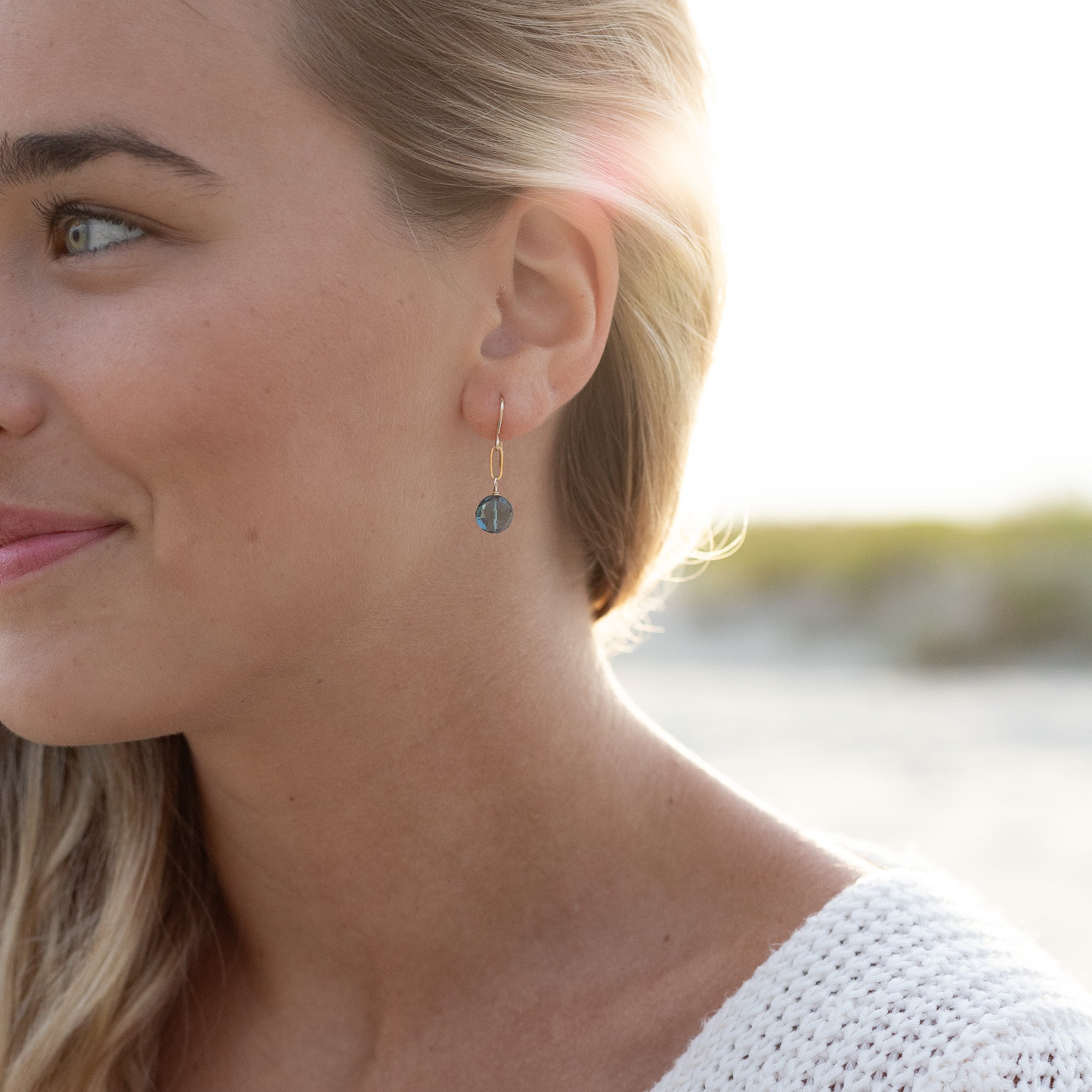 tiny dangle earrings with one gold filled paperclip chain link and one blue crystal on the bottom. Shown in a woman's ear. 