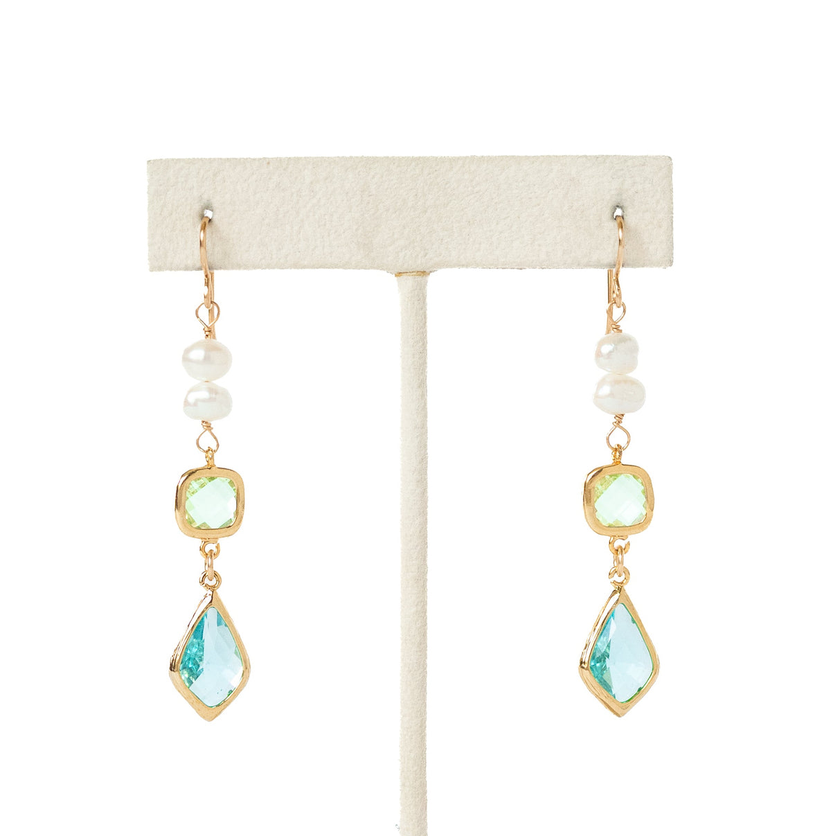A pair of gold drop earrings with white pearl beads, light green oval stones, and light blue teardrop-shaped crystals, displayed on a cream-colored jewelry stand against a white background.