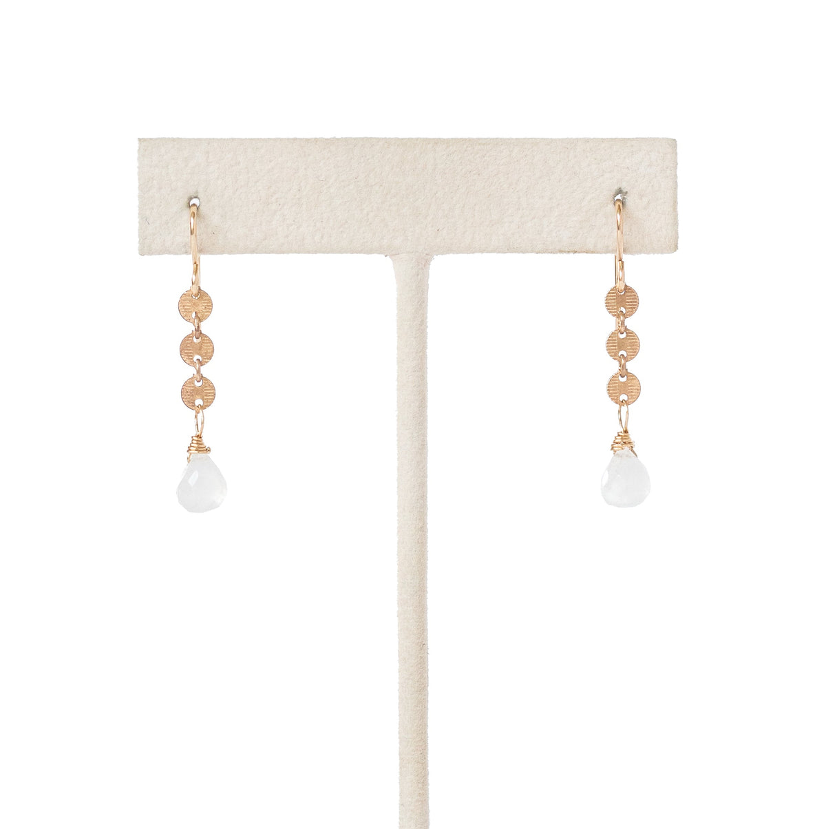 A pair of drop earrings with rose gold disc chain and small teardrop shaped moonstone beads, displayed on a cream-colored jewelry stand against a white background.