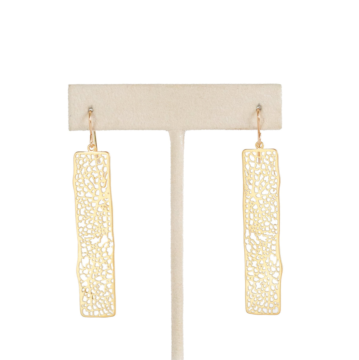 A pair of gold-toned dangle earrings with an intricate openwork filigree pattern, hanging from a cream-colored T-bar jewelry display stand against a solid white background.