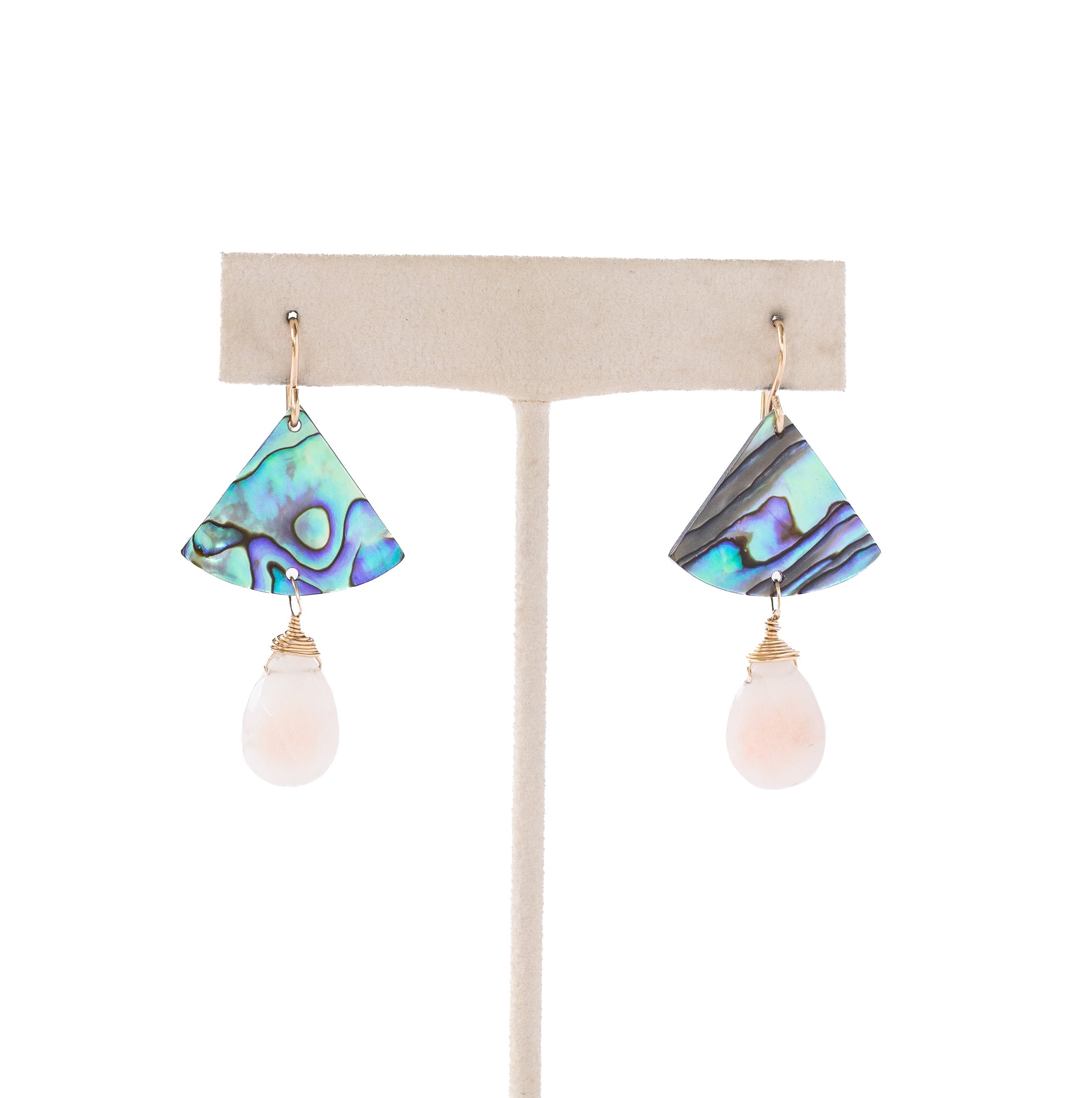 A pair of dangle earrings displayed on a cream-colored jewelry stand against a white background. Each earring features a fan-shaped top made of iridescent abalone shell with blue, green, and purple tones, hanging from a gold post with a pale pink teardrop bead below.