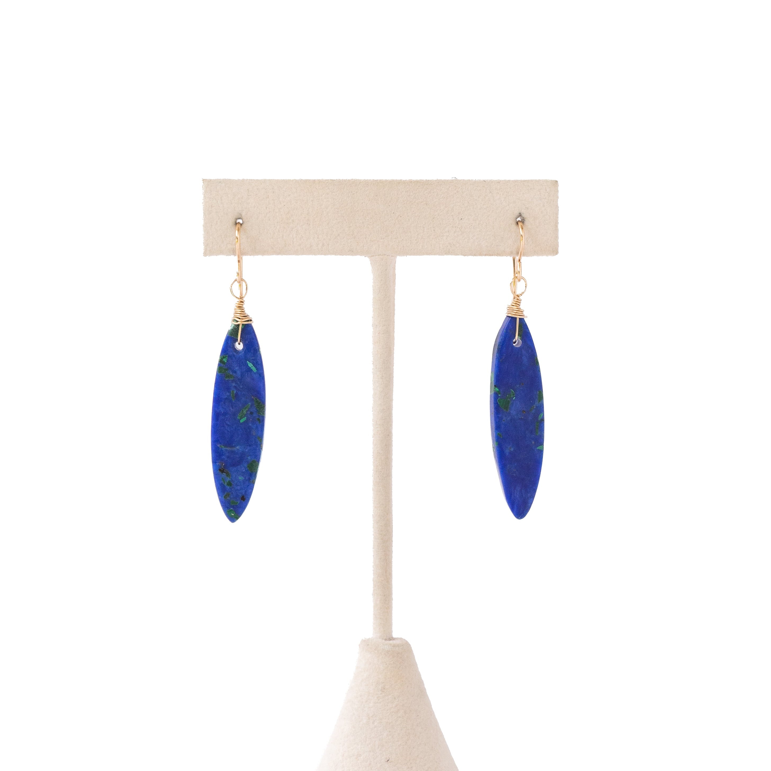 A pair of long oval drop earrings in deep blue lapis lazuli with natural white and green veining, hanging from gold-tone rings on a beige jewelry display stand against a white background.