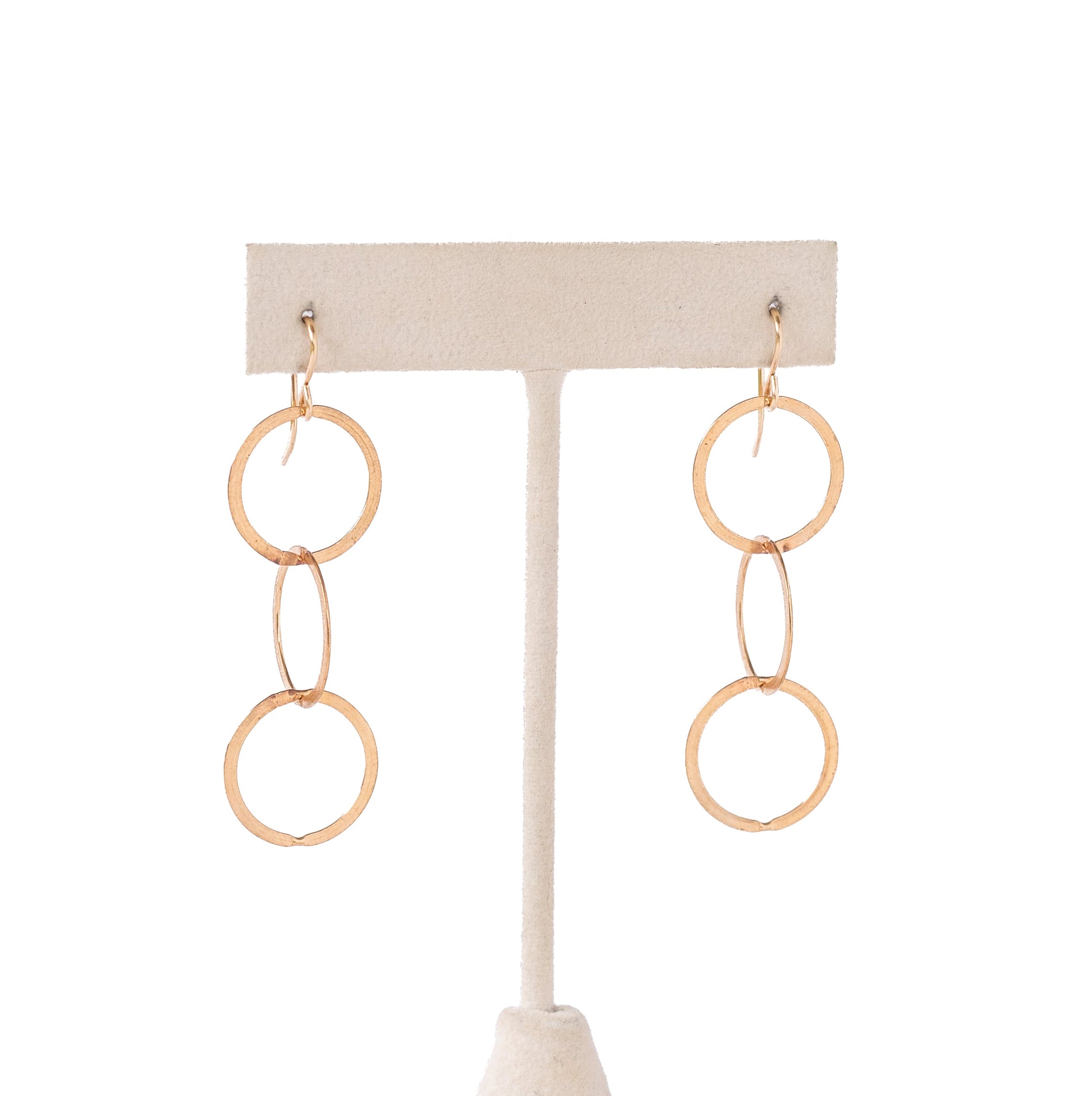 A pair of gold drop earrings with three interlocking circular rings, displayed on a cream-colored fabric jewelry stand against a white background.