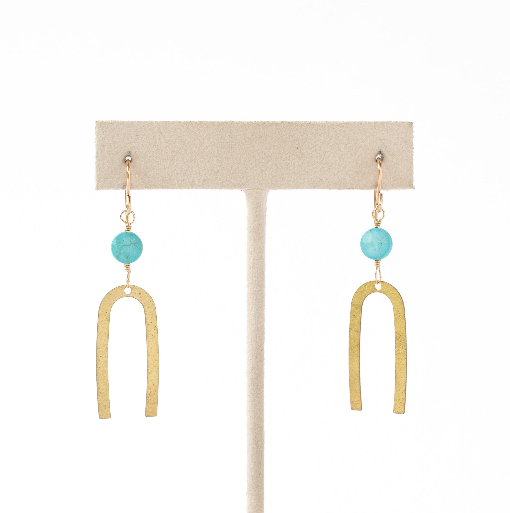 A pair of gold-toned drop earrings with a small round turquoise blue bead and a larger open U-shaped gold pendant, displayed on a cream-colored jewelry stand against a white background.