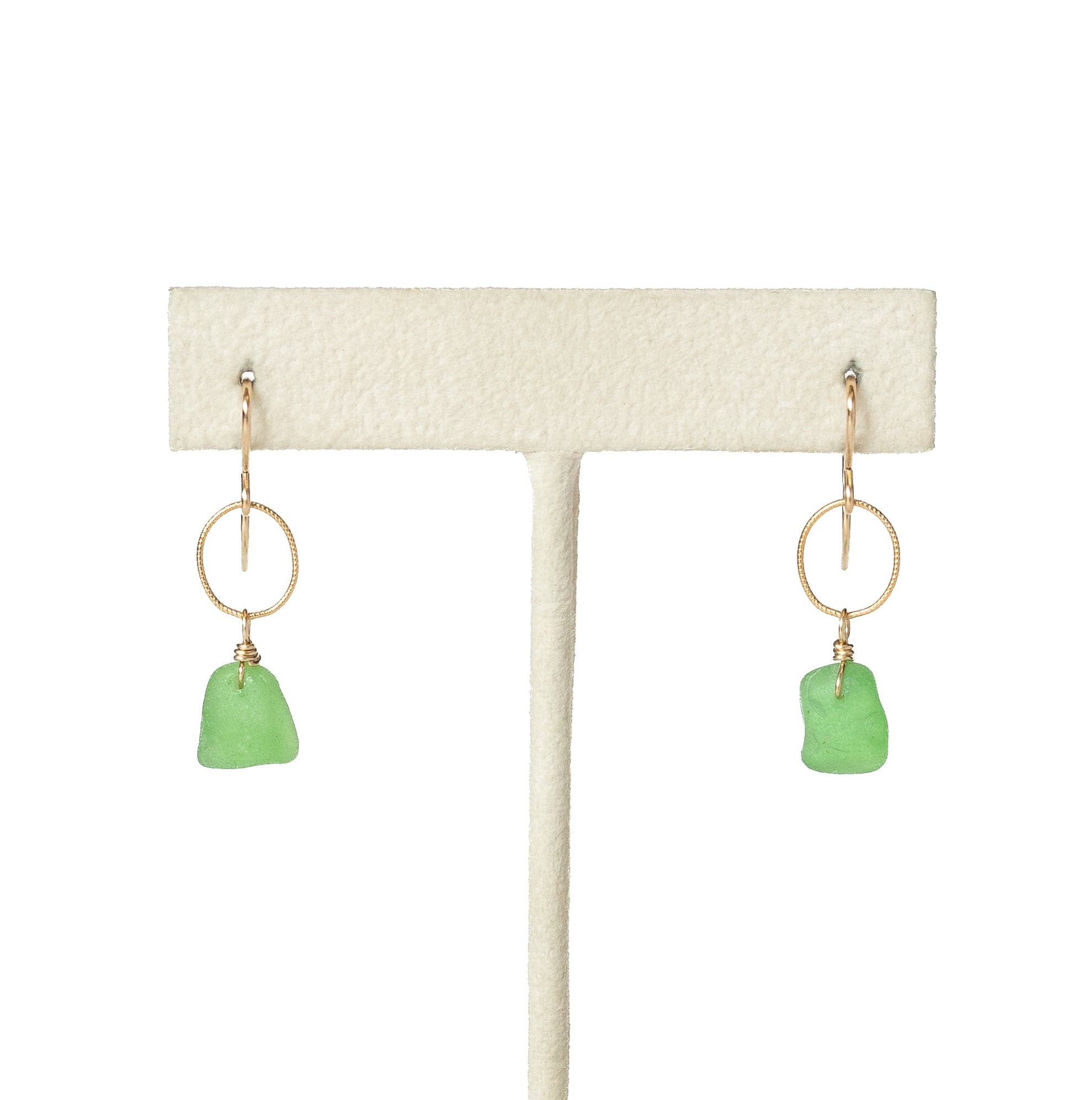 A pair of gold earrings with small green sea glass pendants, displayed on a cream-colored jewelry stand against a white background.