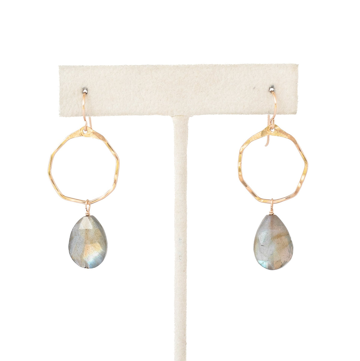 A pair of gold hoop earrings with teardrop-shaped iridescent gray-blue stones, displayed on a cream-colored jewelry stand against a white background.