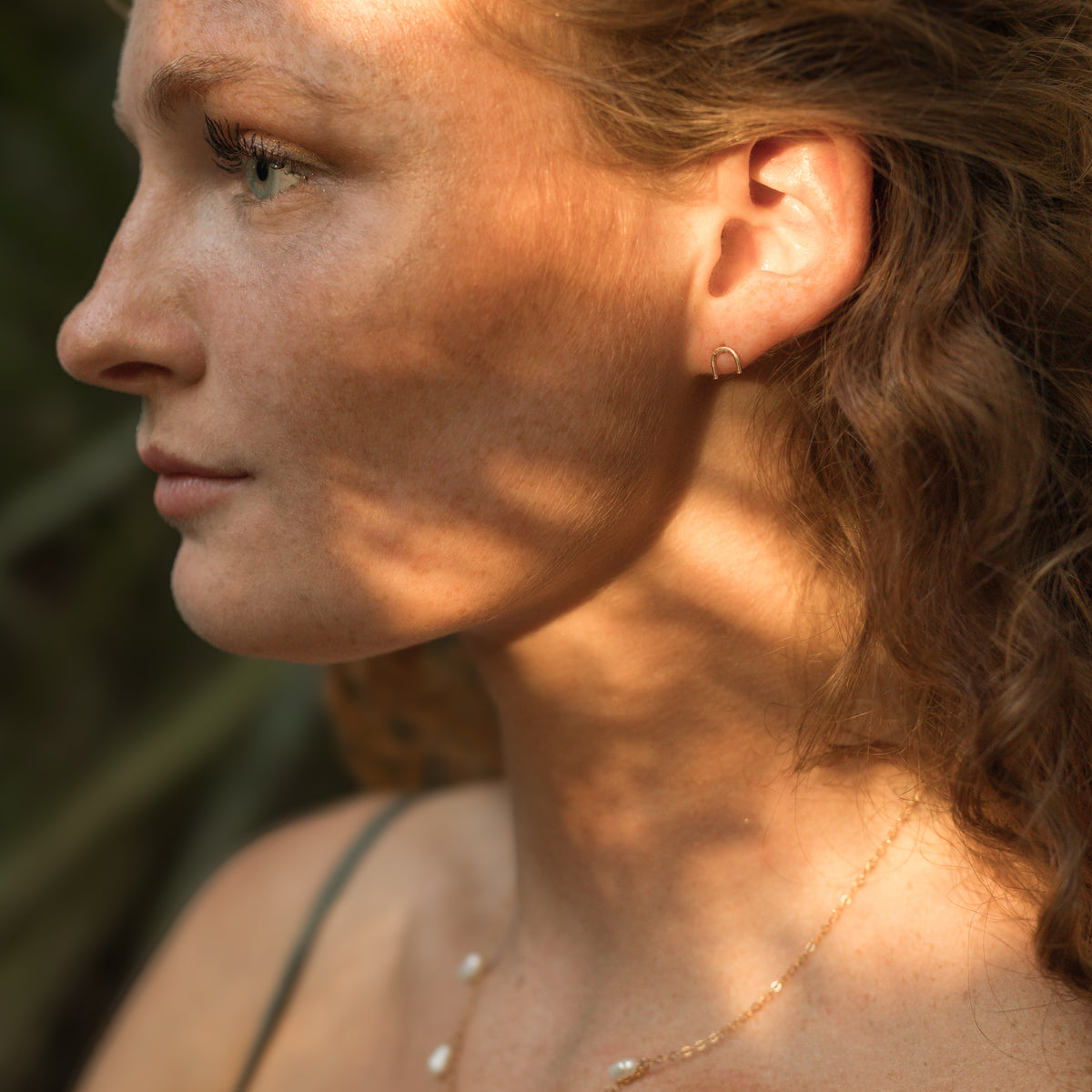 Close-up side profile of a woman outdoors wearing a small gold hoop earring, with warm sunlight and leaf shadows across her face and neck.
