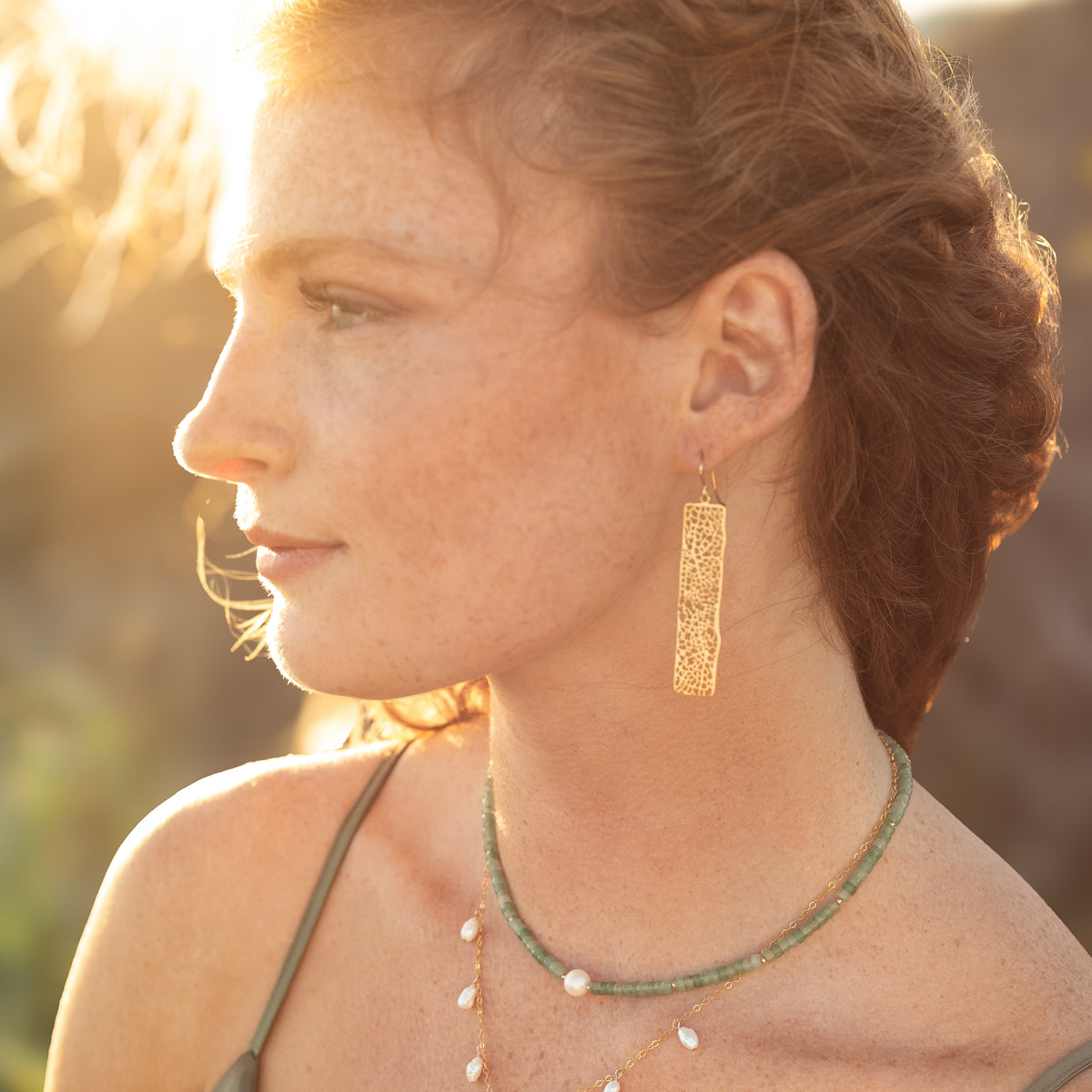 A woman in profile wearing a long, rectangular gold dangle earring with an intricate openwork pattern, styled with a green beaded necklace and pearl necklace against a warm, backlit outdoor background.