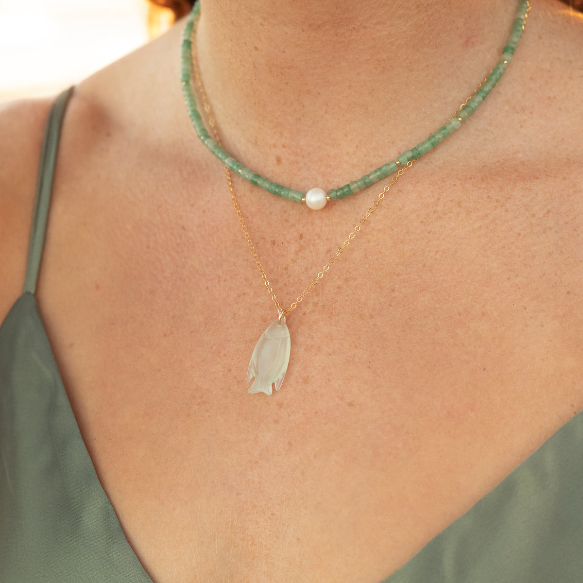 A close-up of a person's neck and chest wearing two layered necklaces: a green beaded choker with a white pearl, and a delicate gold chain with a white leaf-shaped pendant.