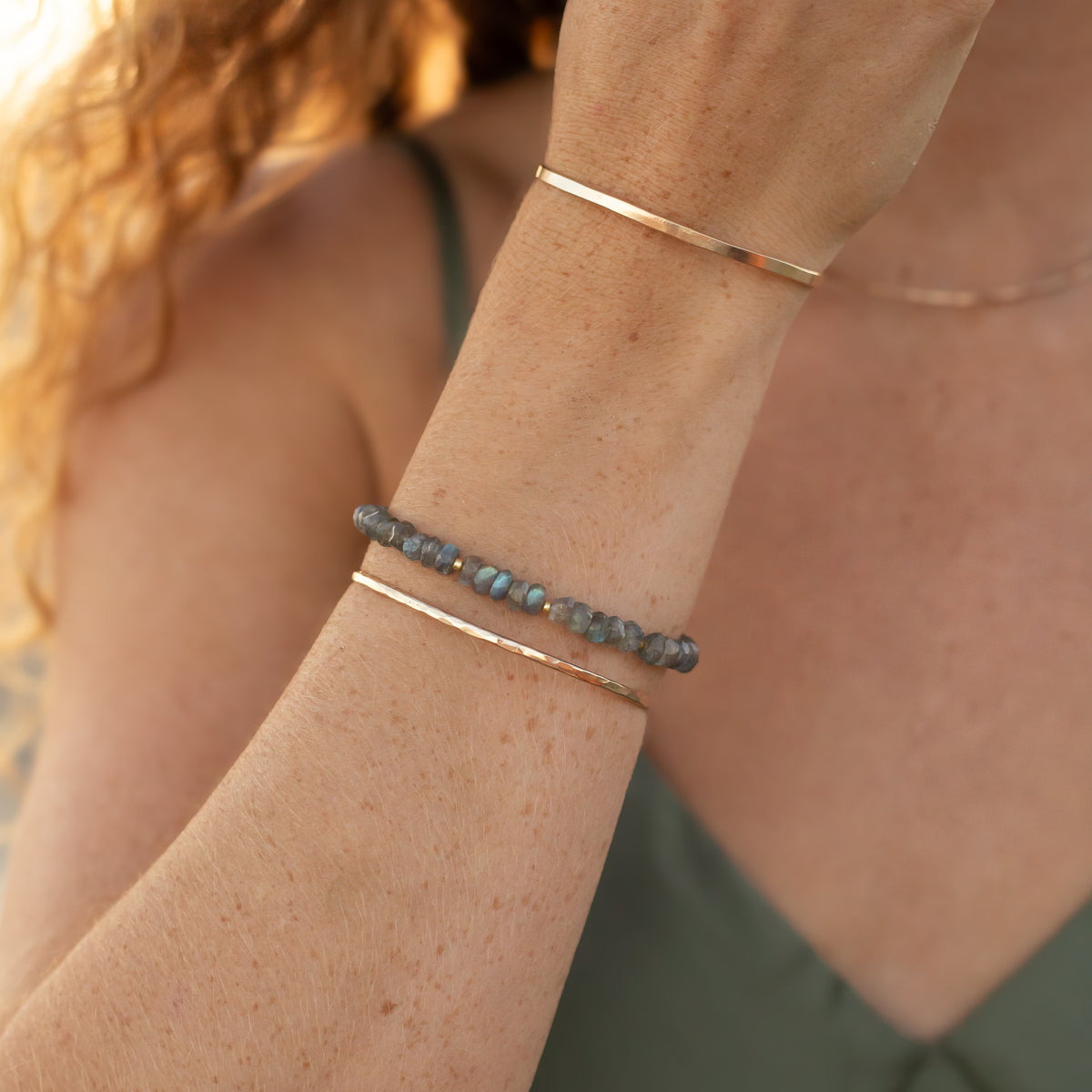 A close-up of a person's wrist wearing three bracelets: two slim gold-tone bangle bracelets and one beaded bracelet with small gray-blue stones, shown against skin and a green top.