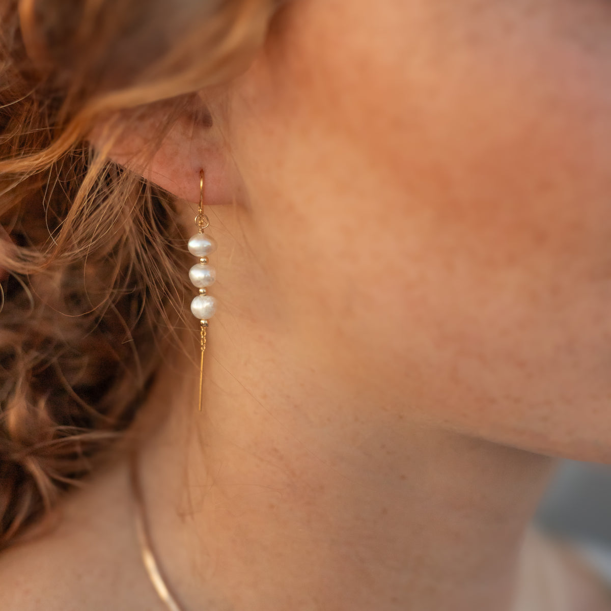 A close-up of a person wearing a delicate gold dangle earring with three small white pearls, shown against skin and hair.