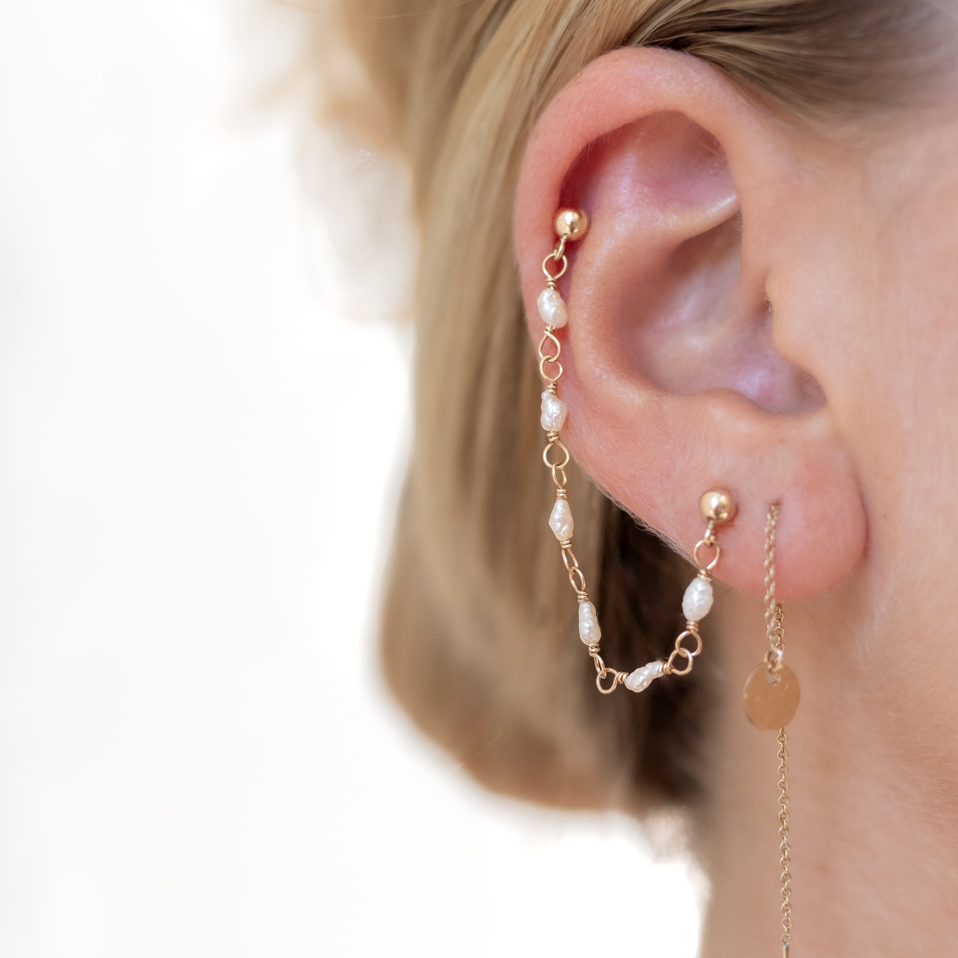 An earring with a gold ball stud in first ear piercing and a gold bar stud in cartilage piercing connected by small rice pearls wire wrapped to each other with gold wire. 