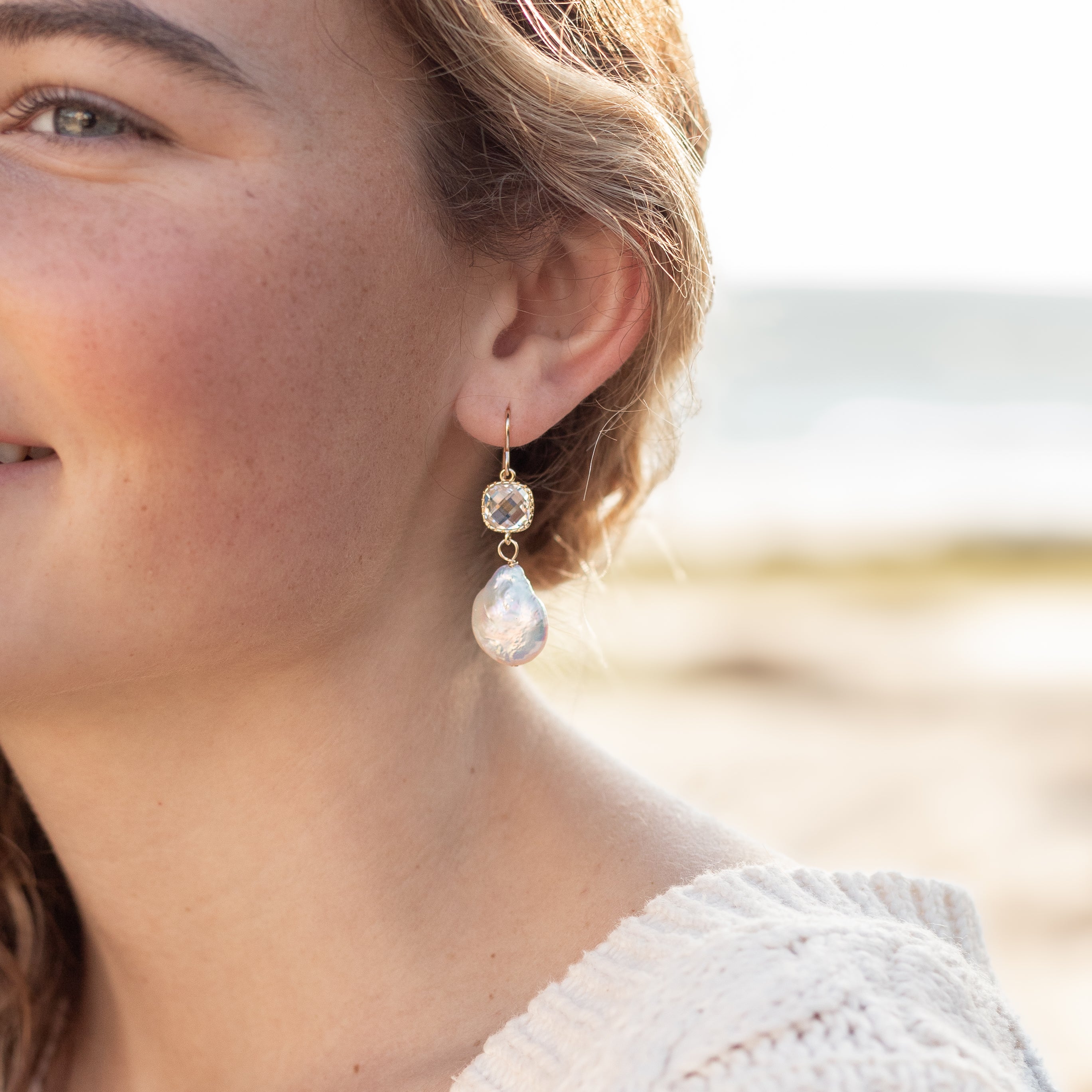 A pair of earrings with crystals wrapped in gold. There is a dime sized pearl hanging from the bottom. Shown dangling from woman’s ear. 