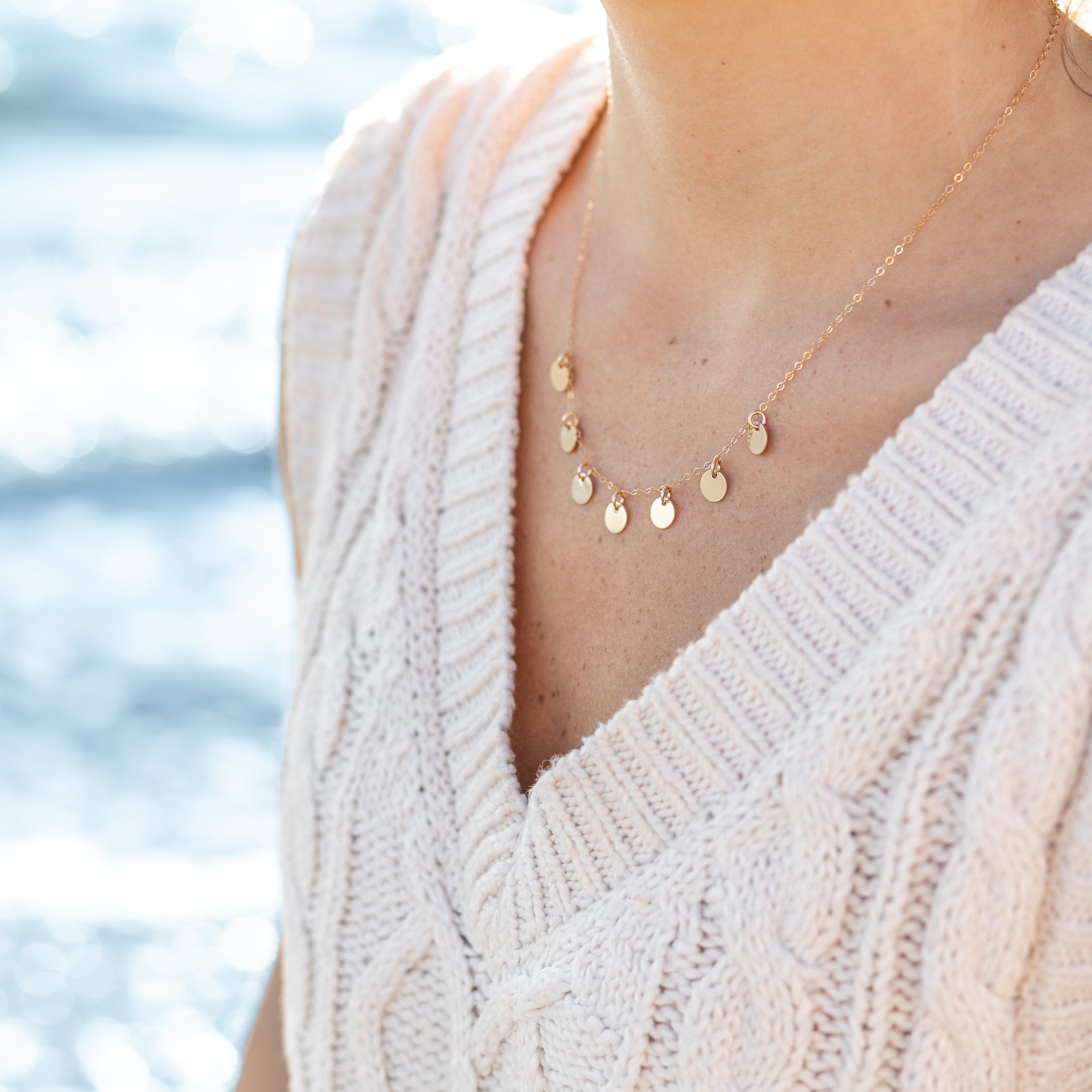 gold necklace with seven tiny circles attached to it. They are about a pea size and spaced about half an inch apart. Shown on a woman’s neck.