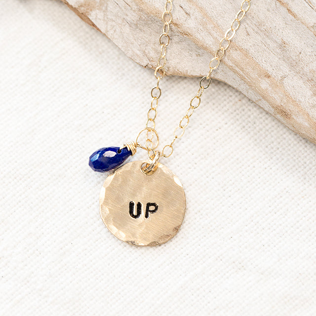 Gold half inch disc with the word UP with a blue lapis stone on a wood background. 