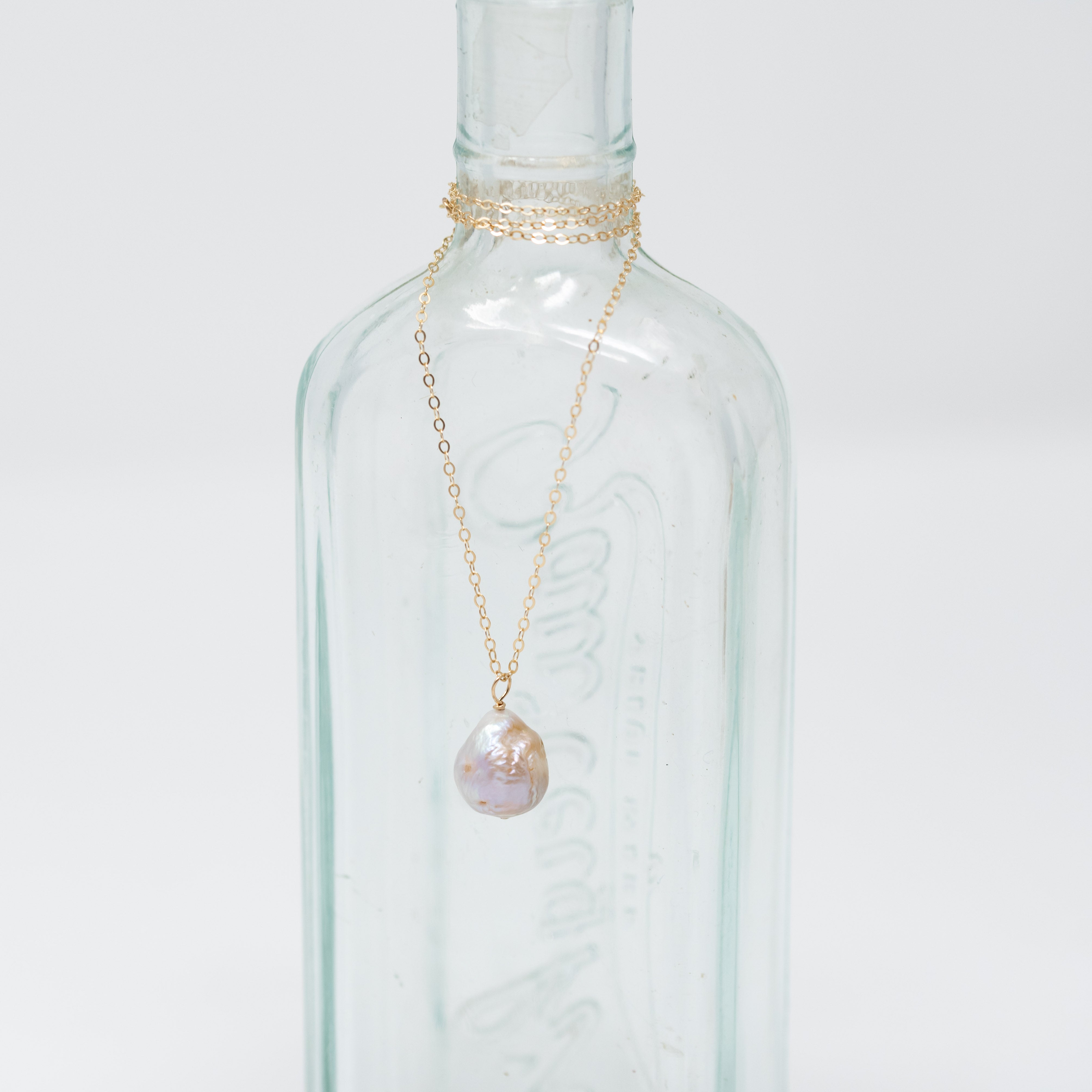 A blush colored pearl around the size of a dime hanging from a gold chain wrapped around a bottle.