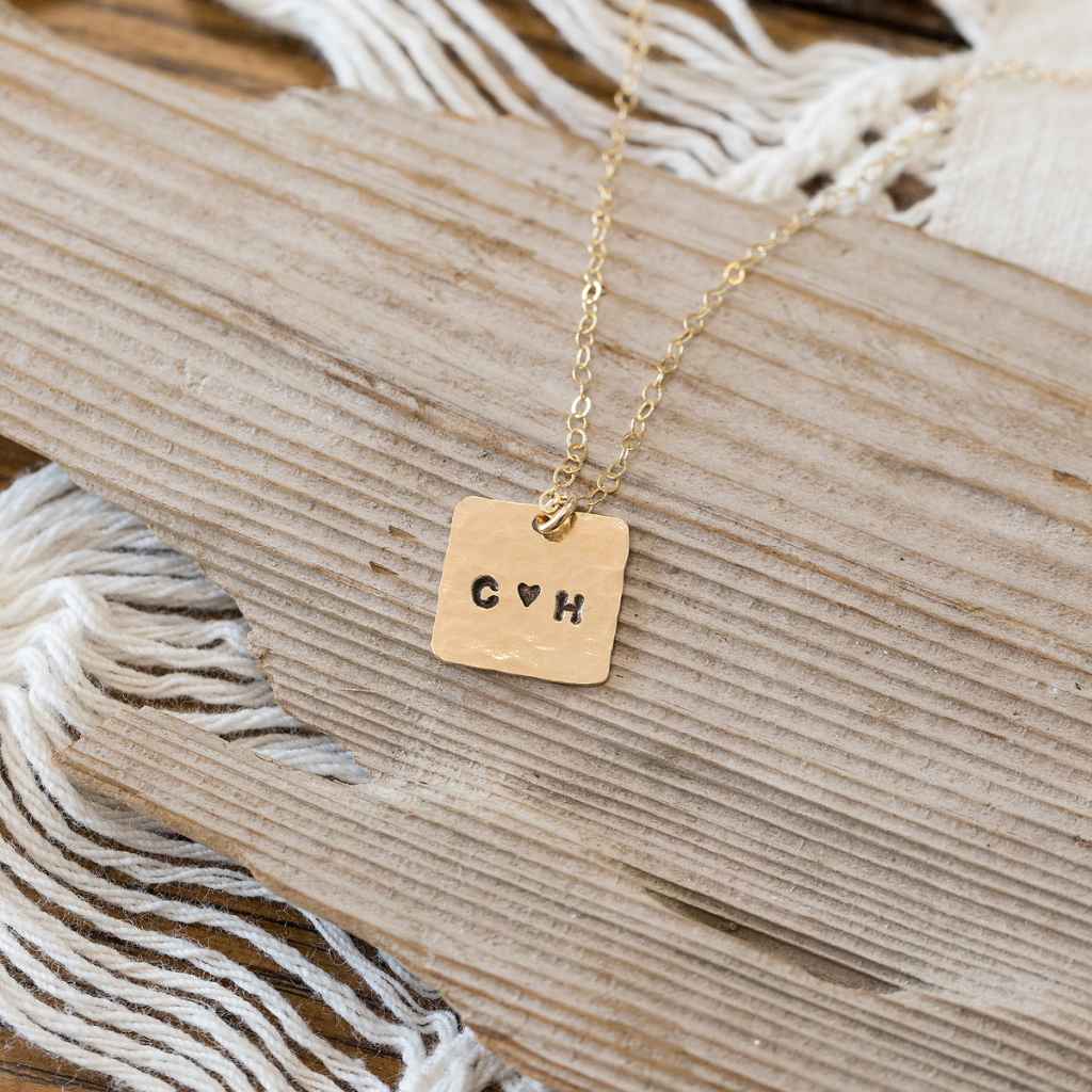 Small dime sized gold square with two initials and a heart in the center. attached to a gold chain laying on a piece of wood.  