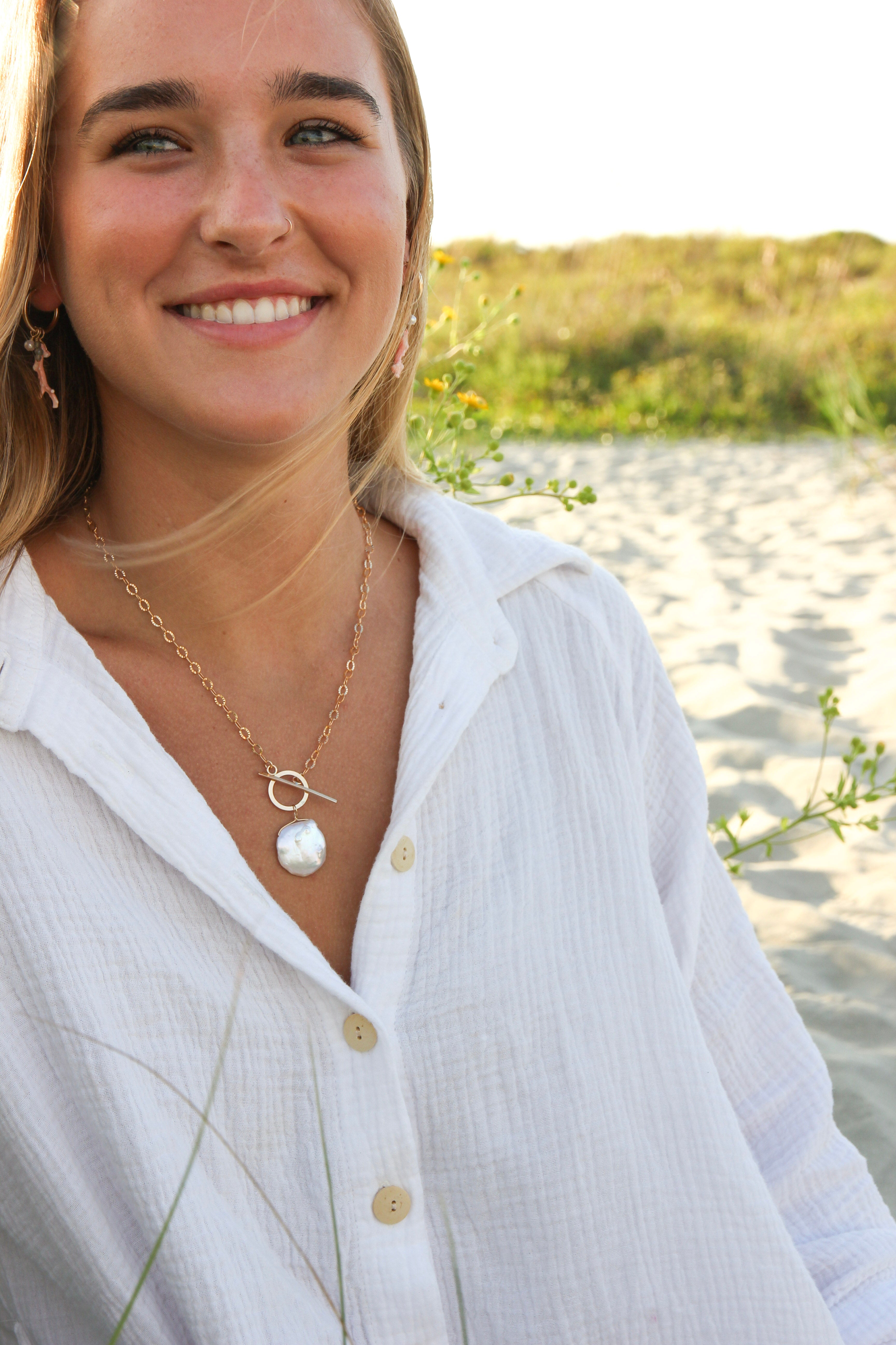 Gold chain necklace with a toggle clasp. There is a dime sized Cornflake pearl wire wrapped on the circular part of the clasp. Shown on a woman's neck who is on the beach.