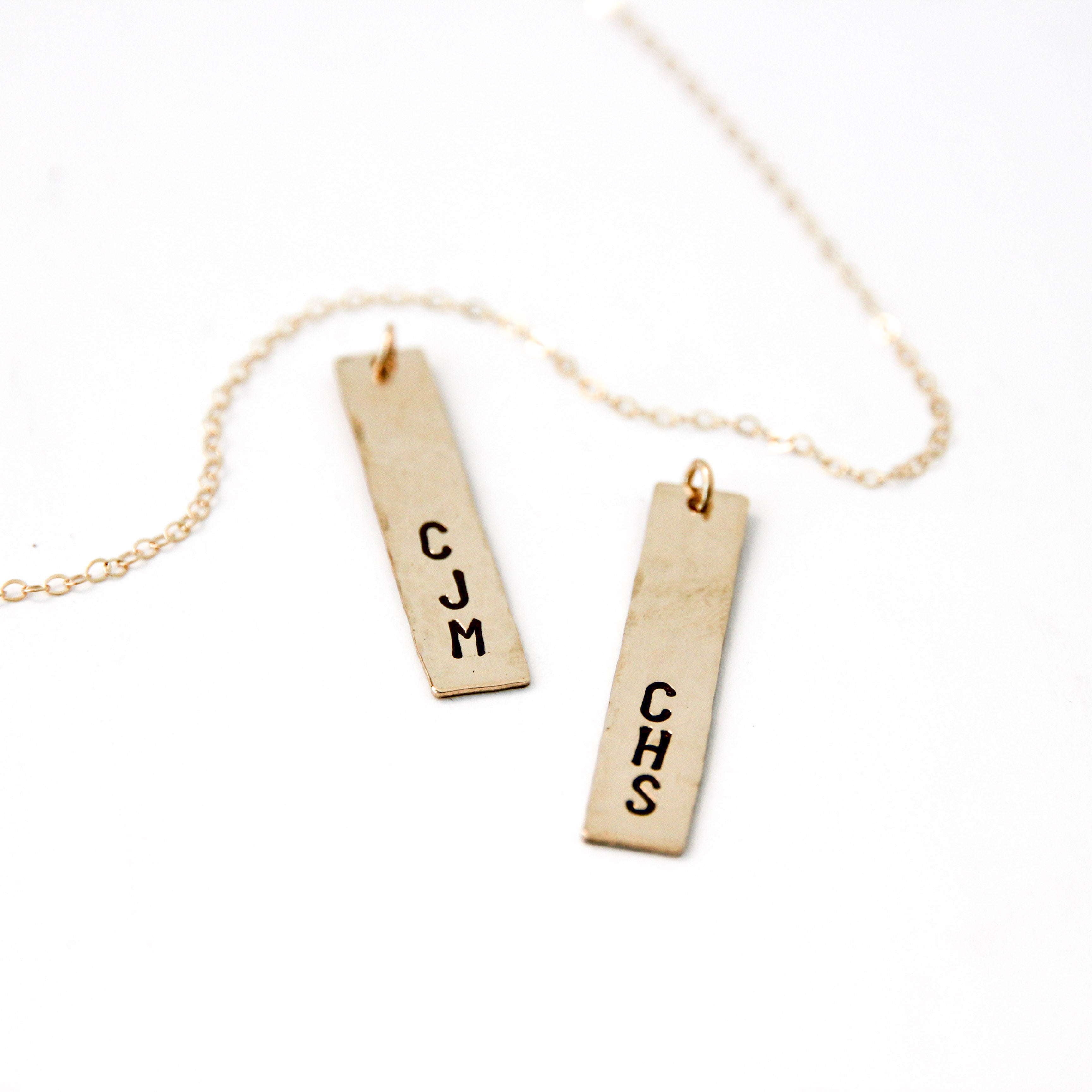 Two vertical gold bar charms with three letter stamped on both of them and a gold chain