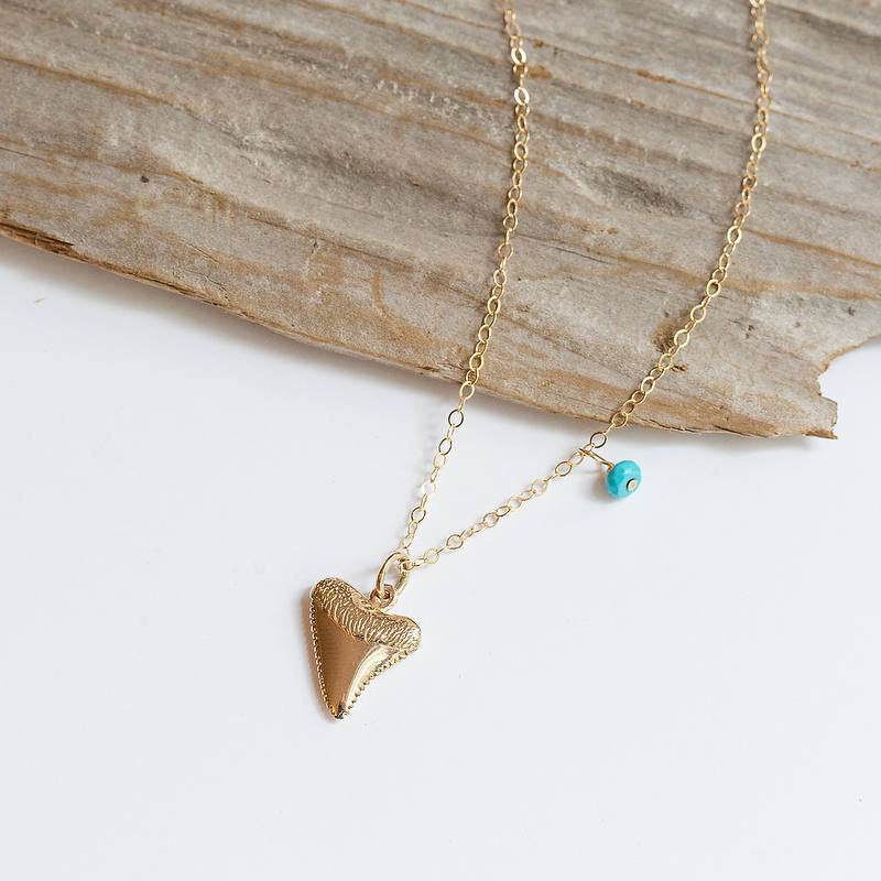 Bronze shark tooth charm hanging from a gold chain. A tiny turquoise bead is hanging with it as well. 